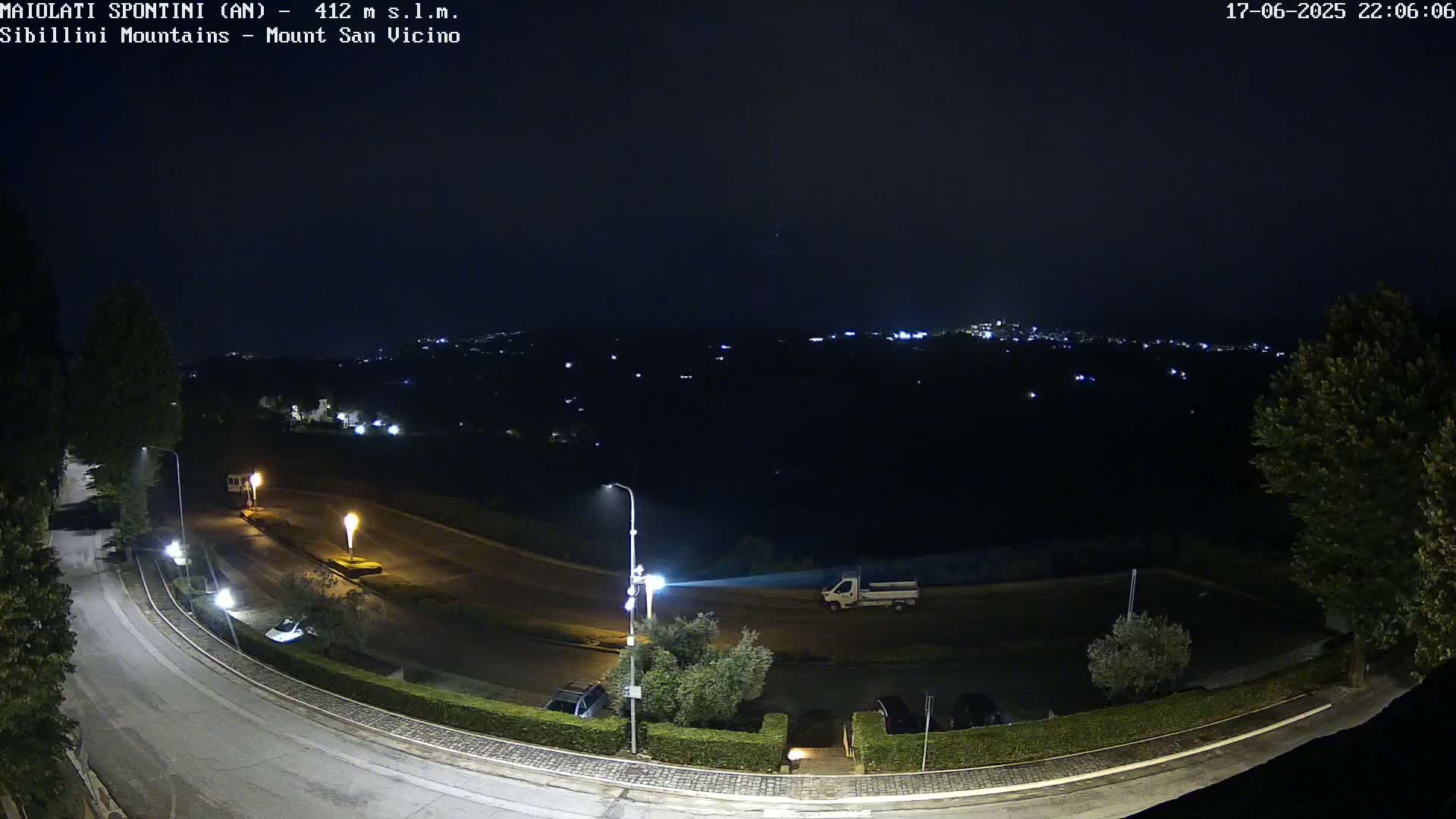 A nighttime view of a roadway curving through a parking lot and toward a hilltop overlooking a distant, dimly lit town under a clear, dark sky.