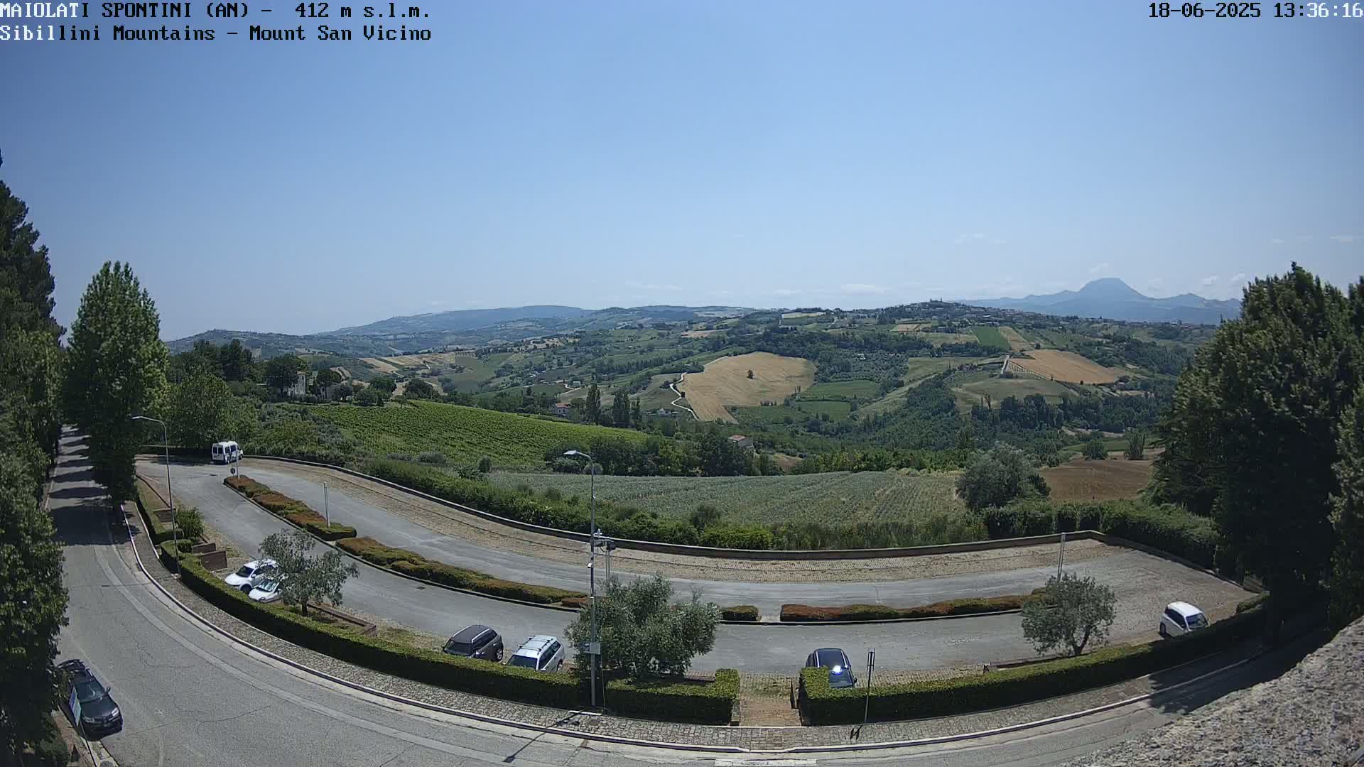 A sunny view of a winding road with parked cars, leading to a parking lot overlooking a vast, hilly landscape of farmland and vineyards under a clear blue sky.