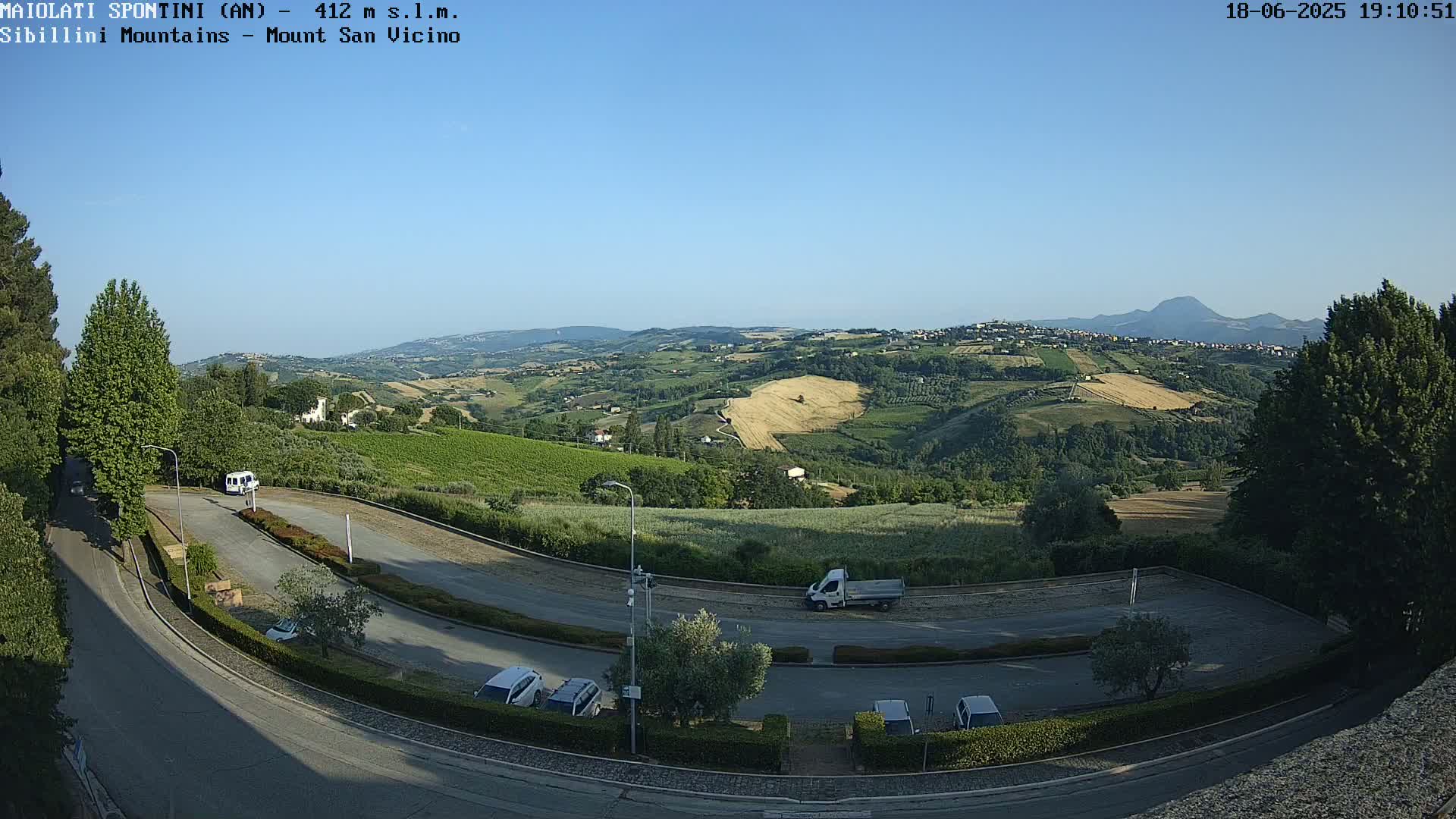 A high-angle, long shot shows a hilly landscape under a clear blue sky, with a parking lot and road in the foreground.
