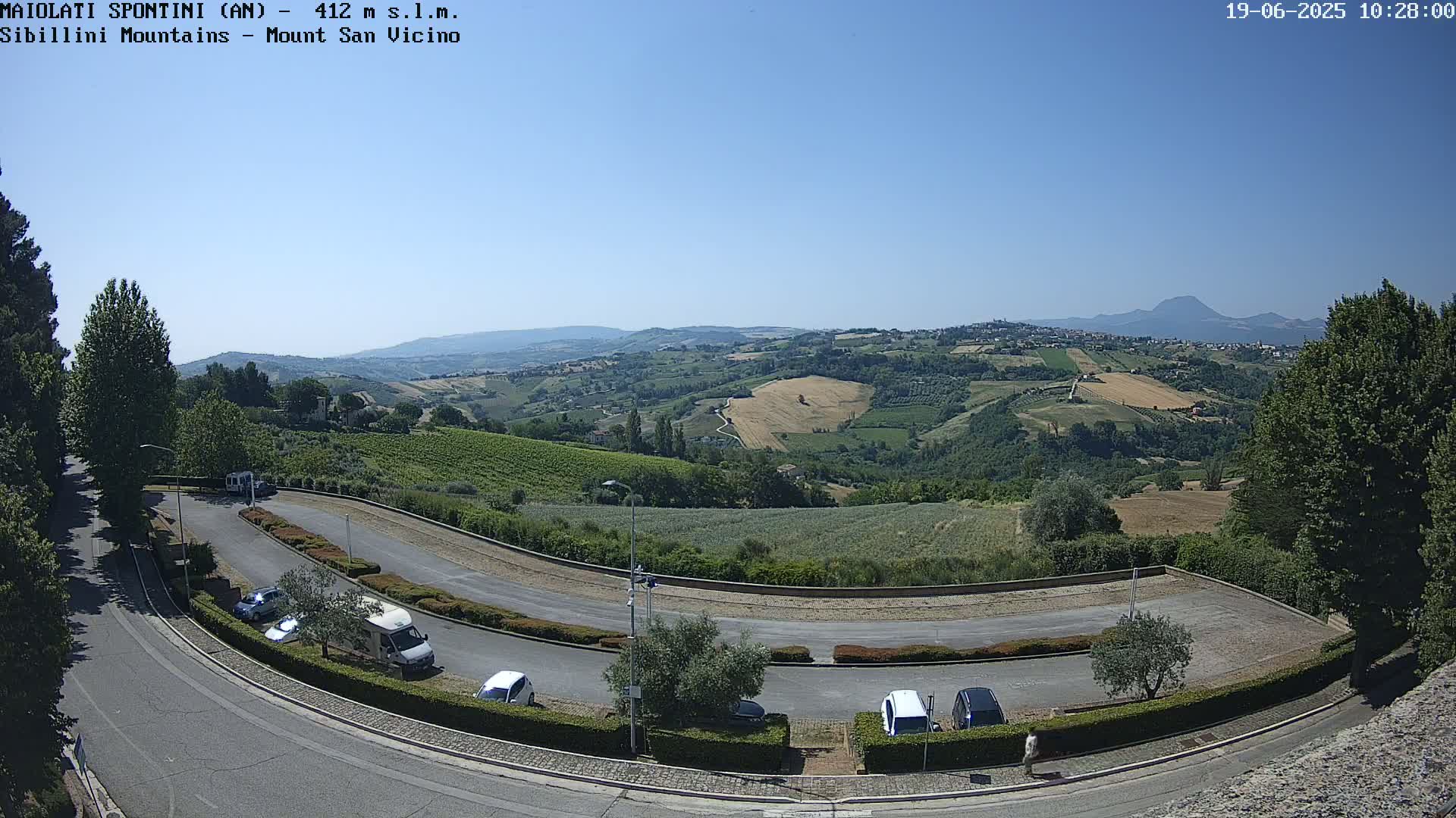 A curved road with parked cars overlooks a sun-drenched valley of rolling hills, vineyards, and farmland under a clear blue sky.