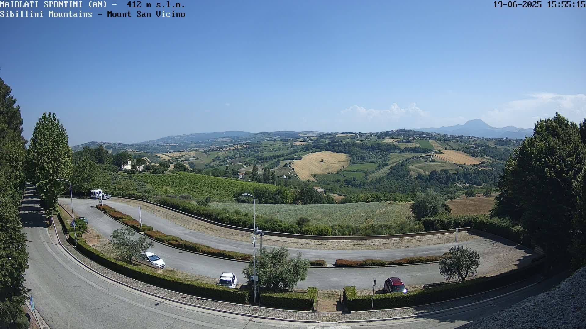 A sunny, clear day overlooks a winding road in a hilly landscape with several parked cars, and distant mountains visible on the horizon.