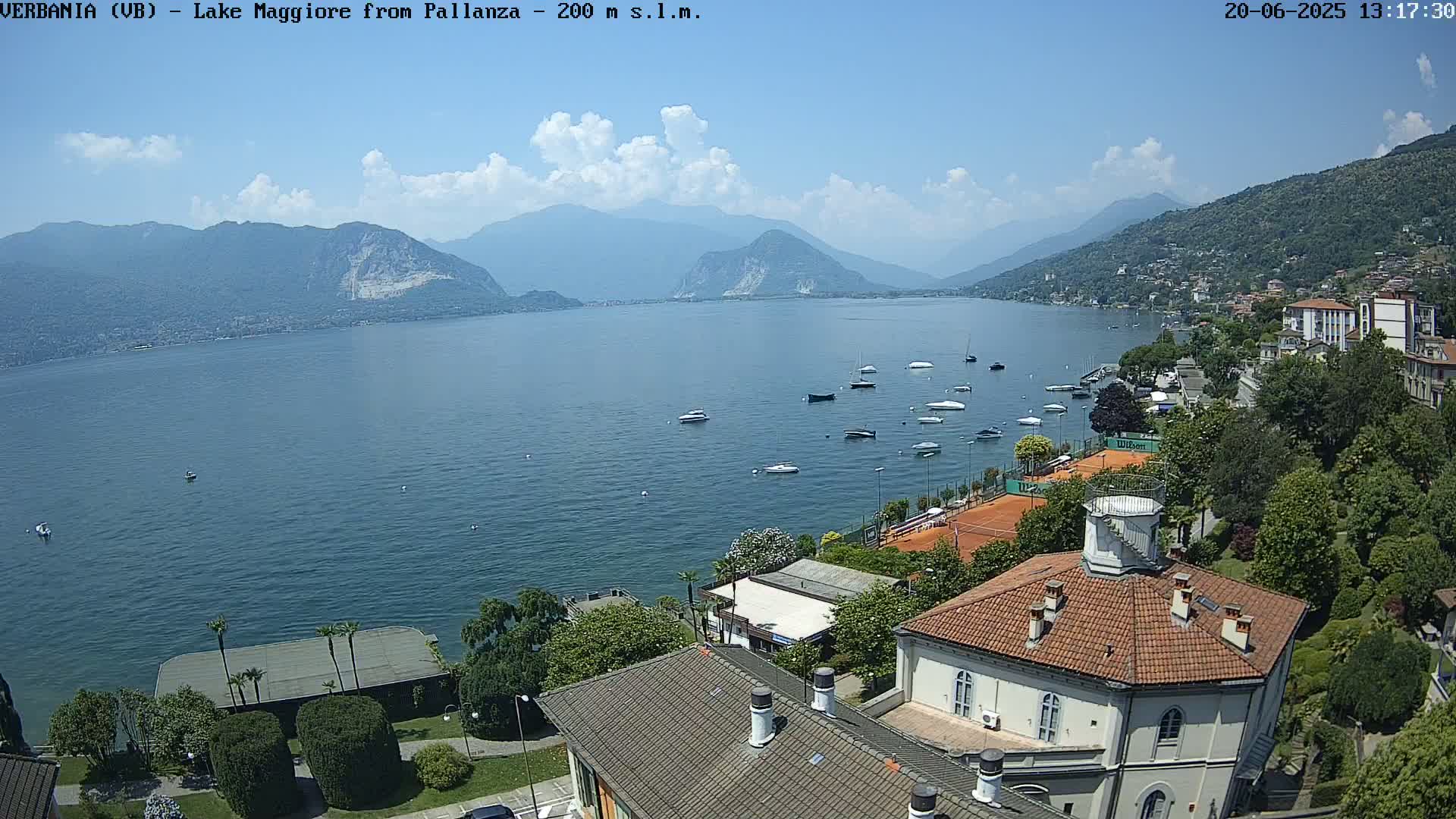 A sunny day overlooking a lake with numerous boats, tennis courts, and buildings nestled along the shoreline, backed by mountains.