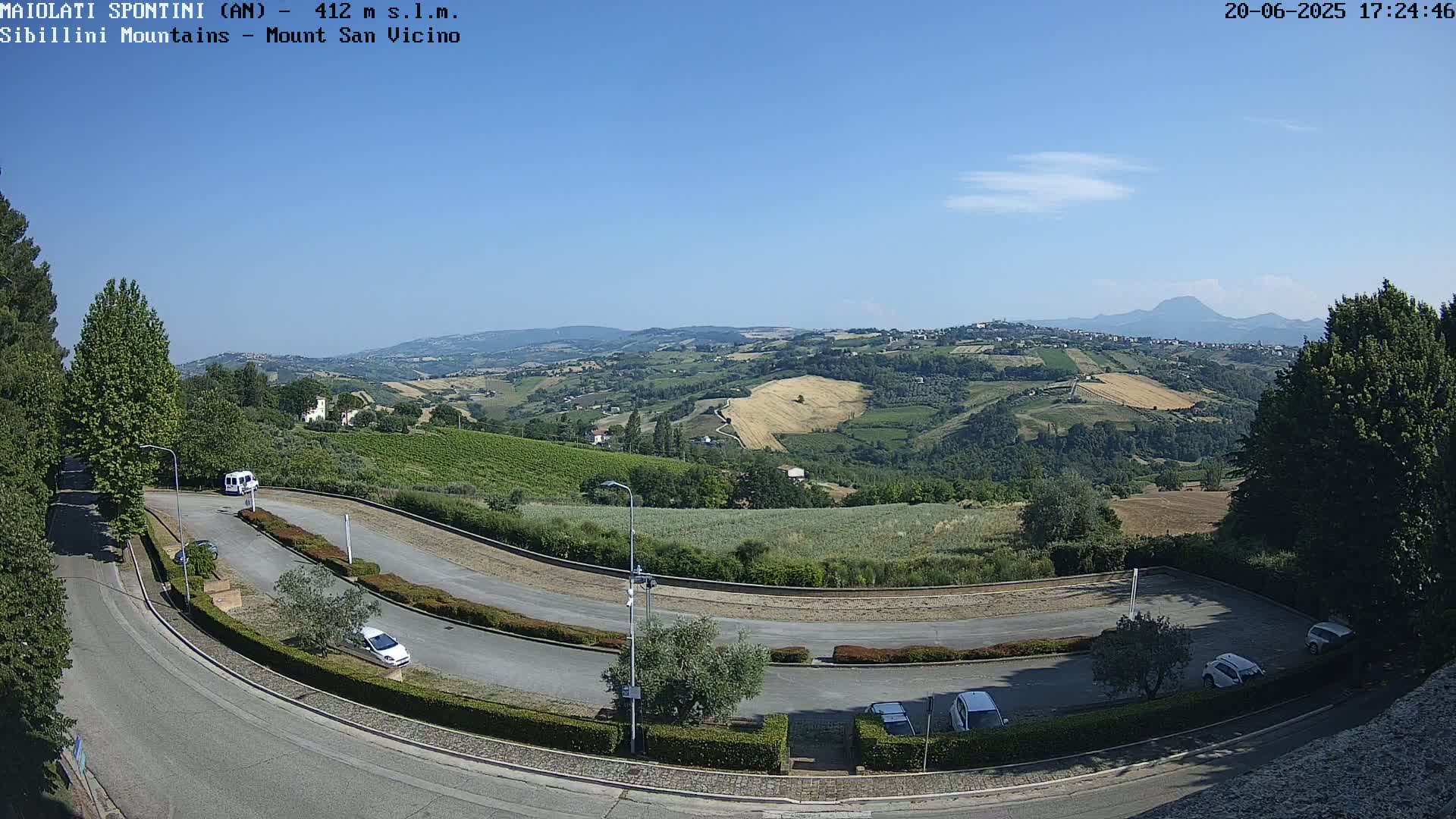 A winding road curves through a parking lot overlooking a sun-drenched valley of green hills and fields under a clear blue sky.