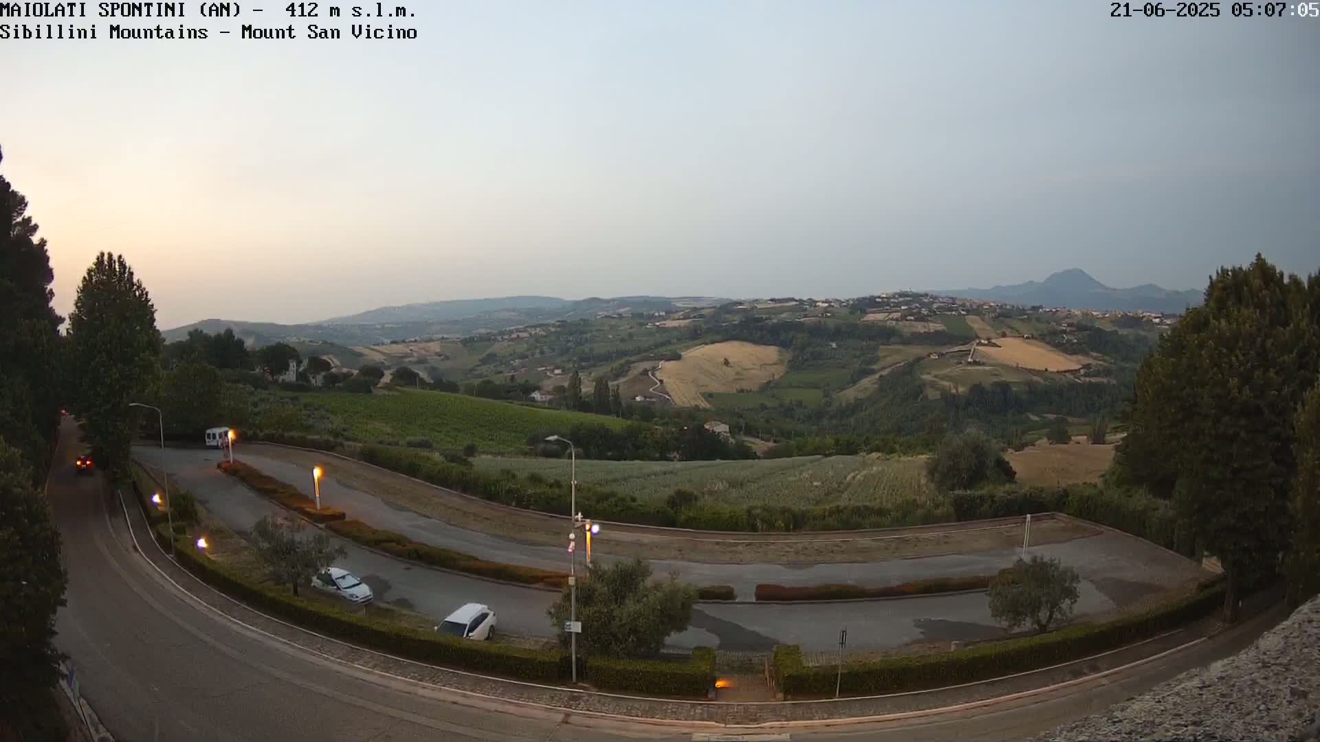 A high-angle, twilight view shows a curving road with parked cars in a parking area, overlooking rolling hills and distant mountains under a hazy sky.