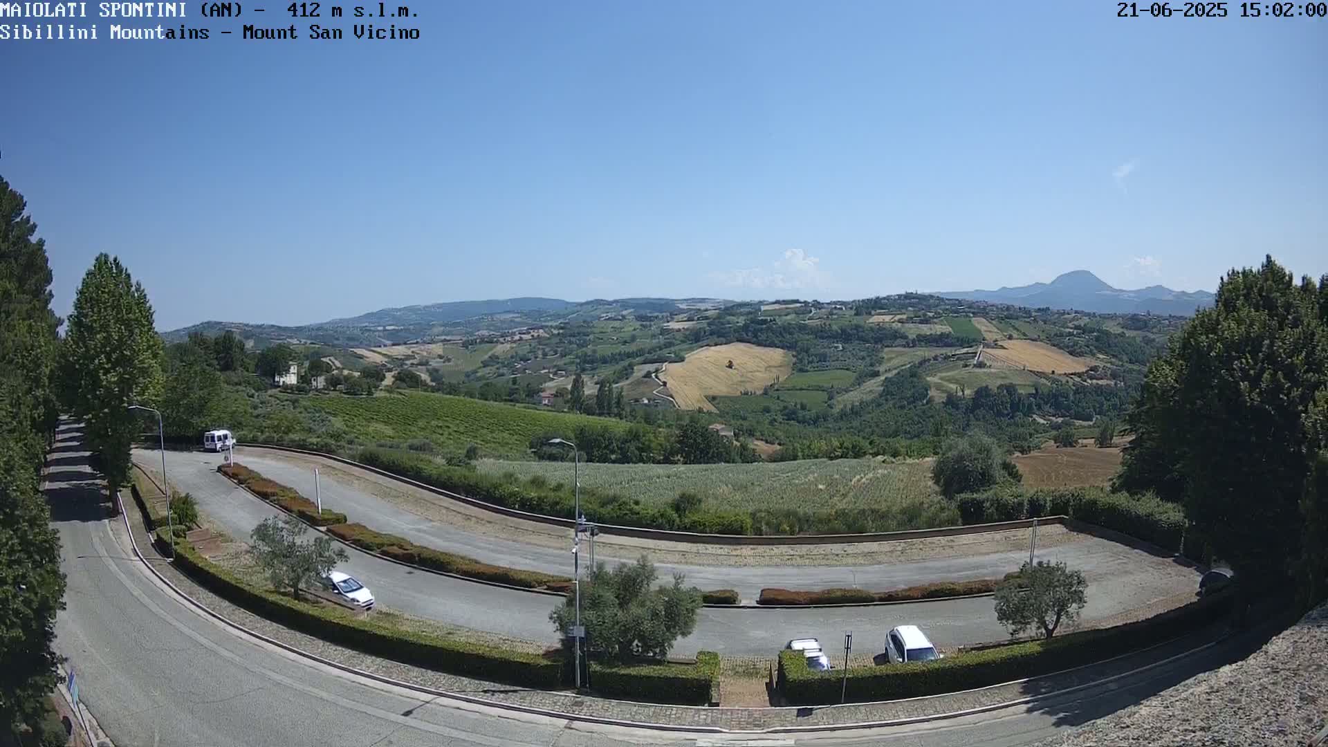 A sunny view of a winding road, several parked cars, and a hilly landscape with fields and trees under a clear blue sky.