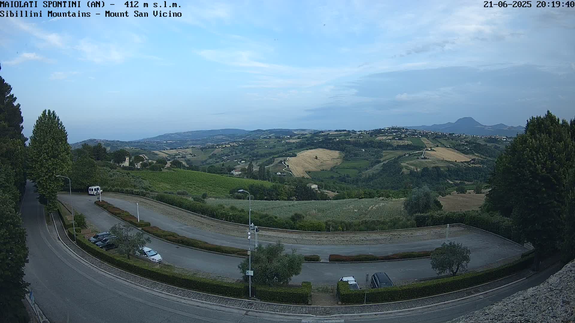 A winding road curves through a parking lot overlooking a hilly landscape under a partly cloudy sky.