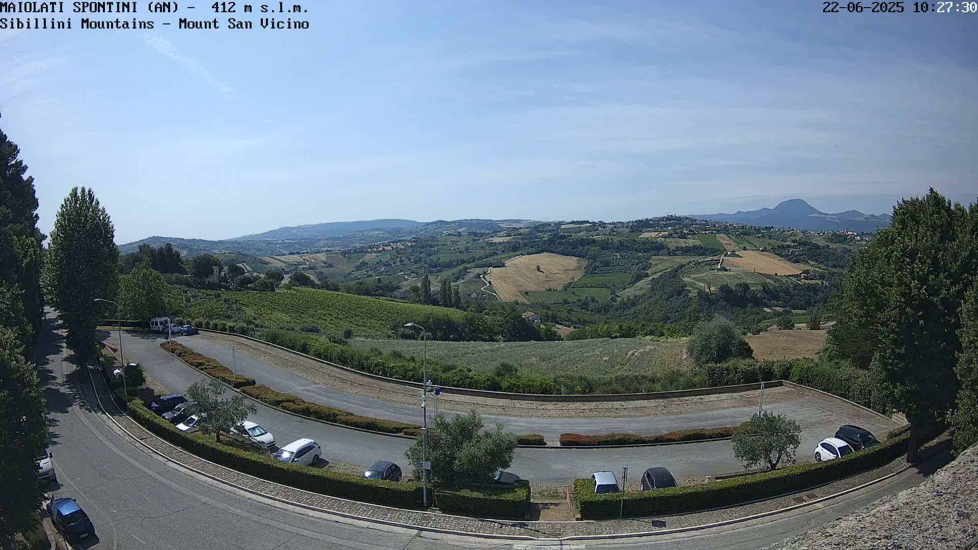 A paved parking lot curves around a hillside overlooking a sunny, rural landscape with fields and distant mountains.