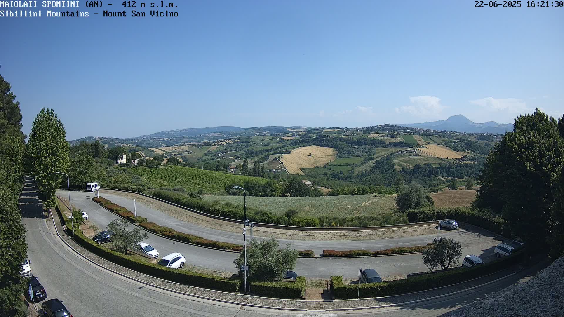 A winding road curves through a parking lot overlooking a sunny, hilly landscape with vineyards and distant mountains under a clear blue sky.