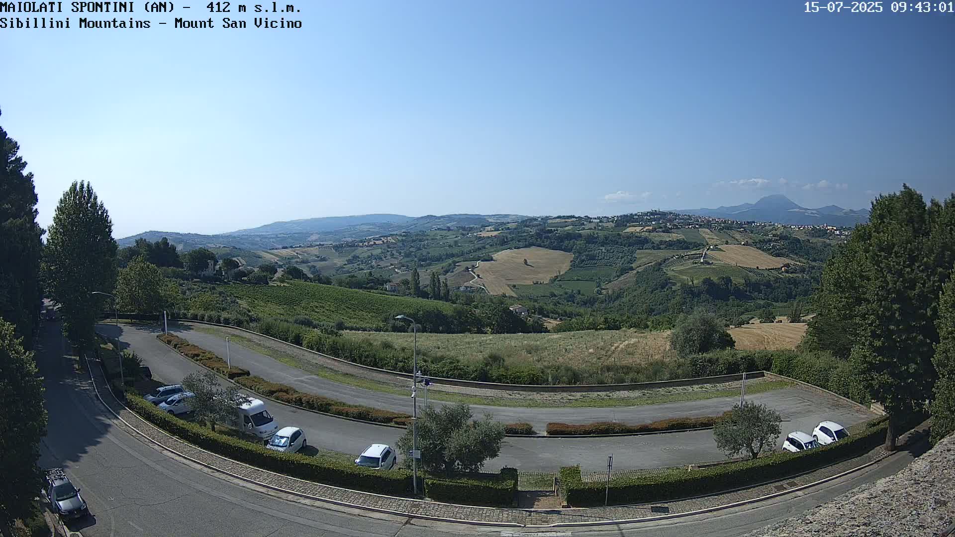 A sunny, clear day overlooks a winding road and parking lot nestled among rolling green hills and vineyards, with distant mountains in the background.