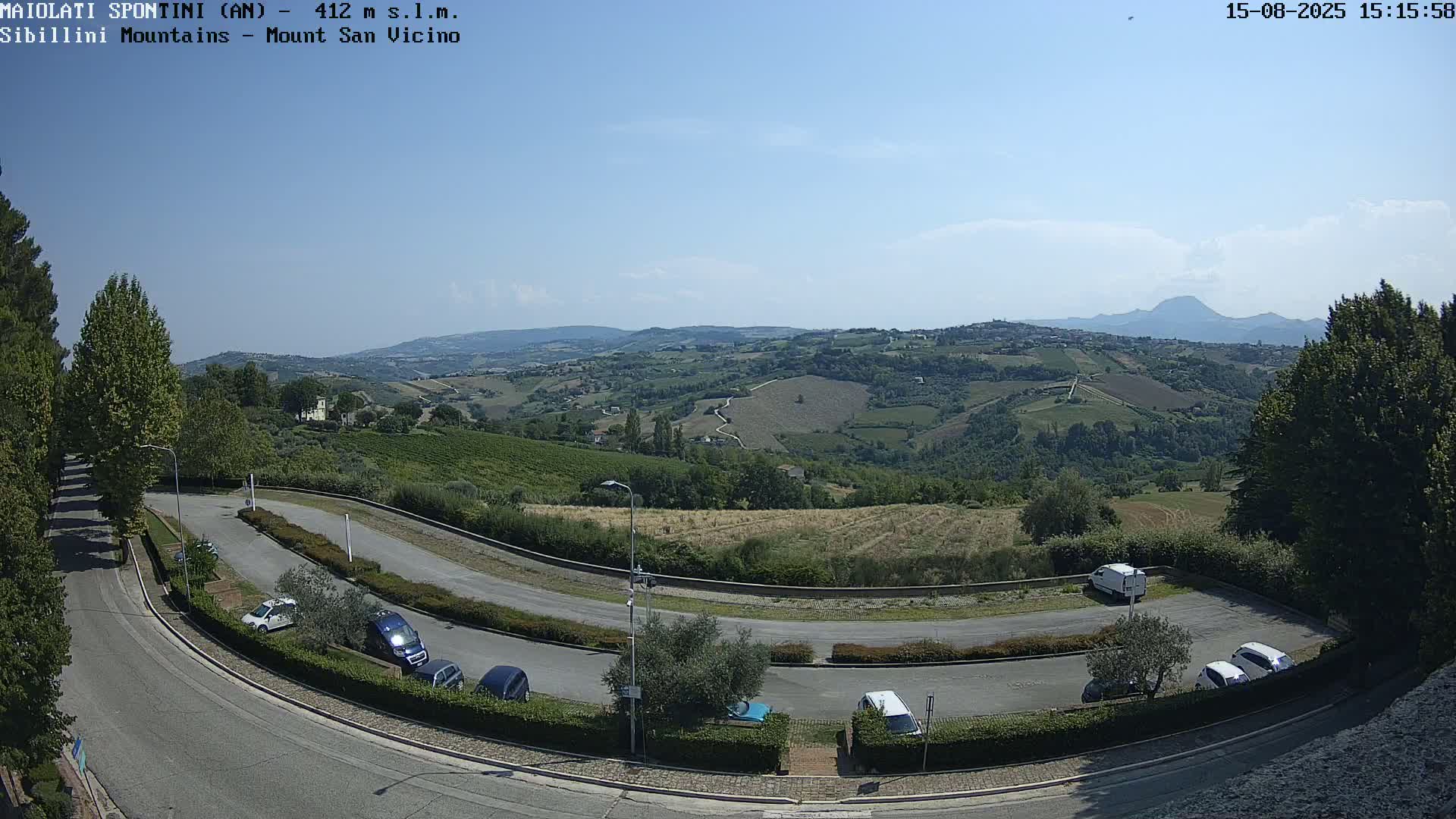 A parking lot with several cars is situated next to a curving road overlooking a sunlit hilly landscape with vineyards and distant mountains under a clear sky.