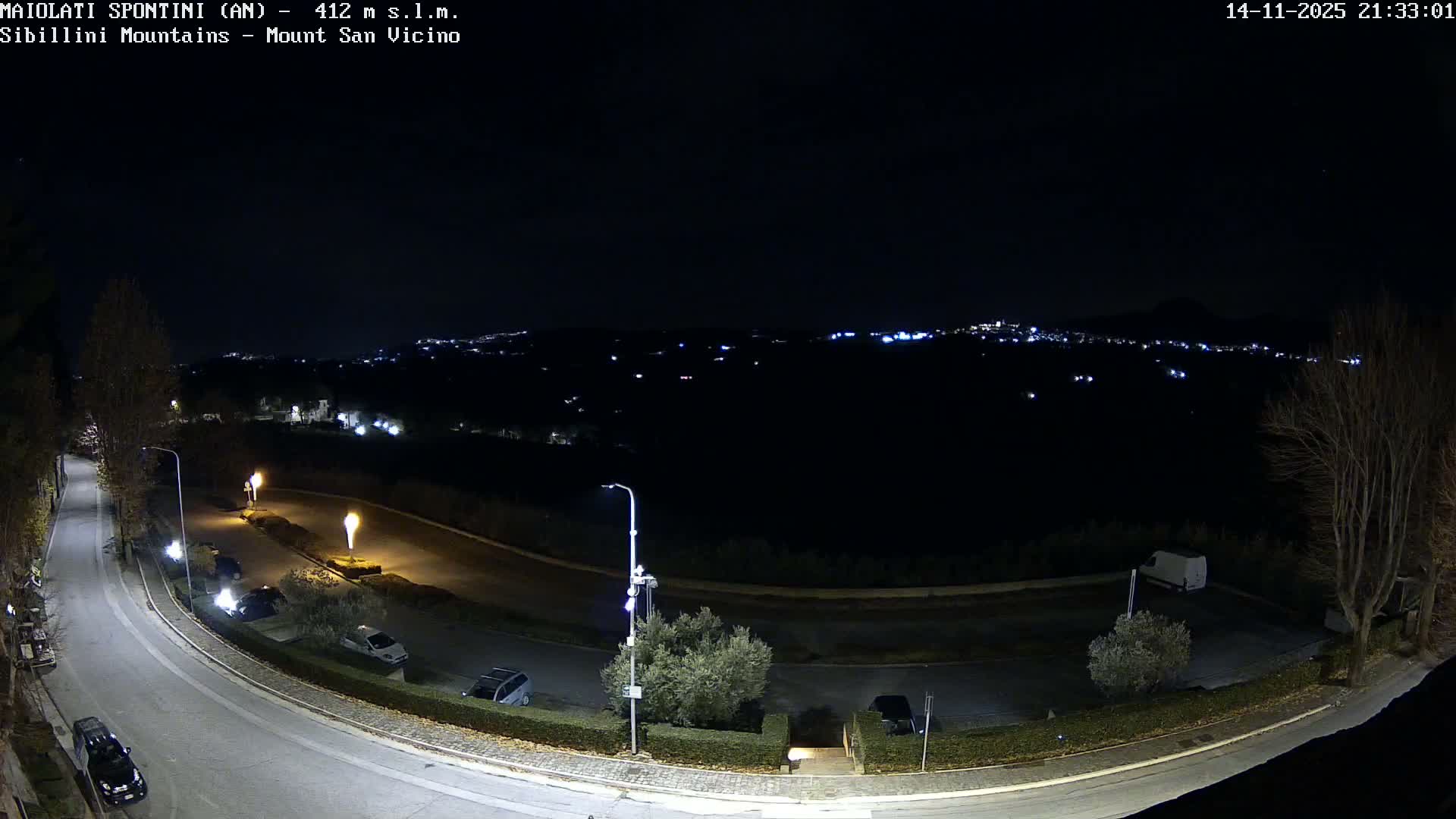 A clear night reveals a winding road illuminated by streetlights, with parked cars and scattered trees, overlooking a dark valley dotted with distant city lights and a faint mountain silhouette under a clear sky.