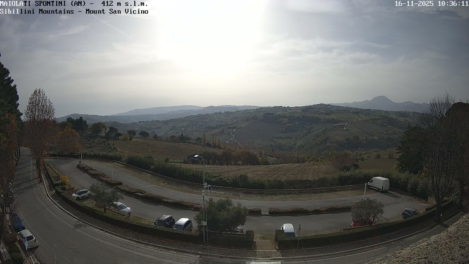 A wide-angle view captures a winding road with parked cars in the foreground, leading into a vast landscape of rolling, autumnal hills with fields, scattered trees, and distant hazy mountains under an overcast sky.