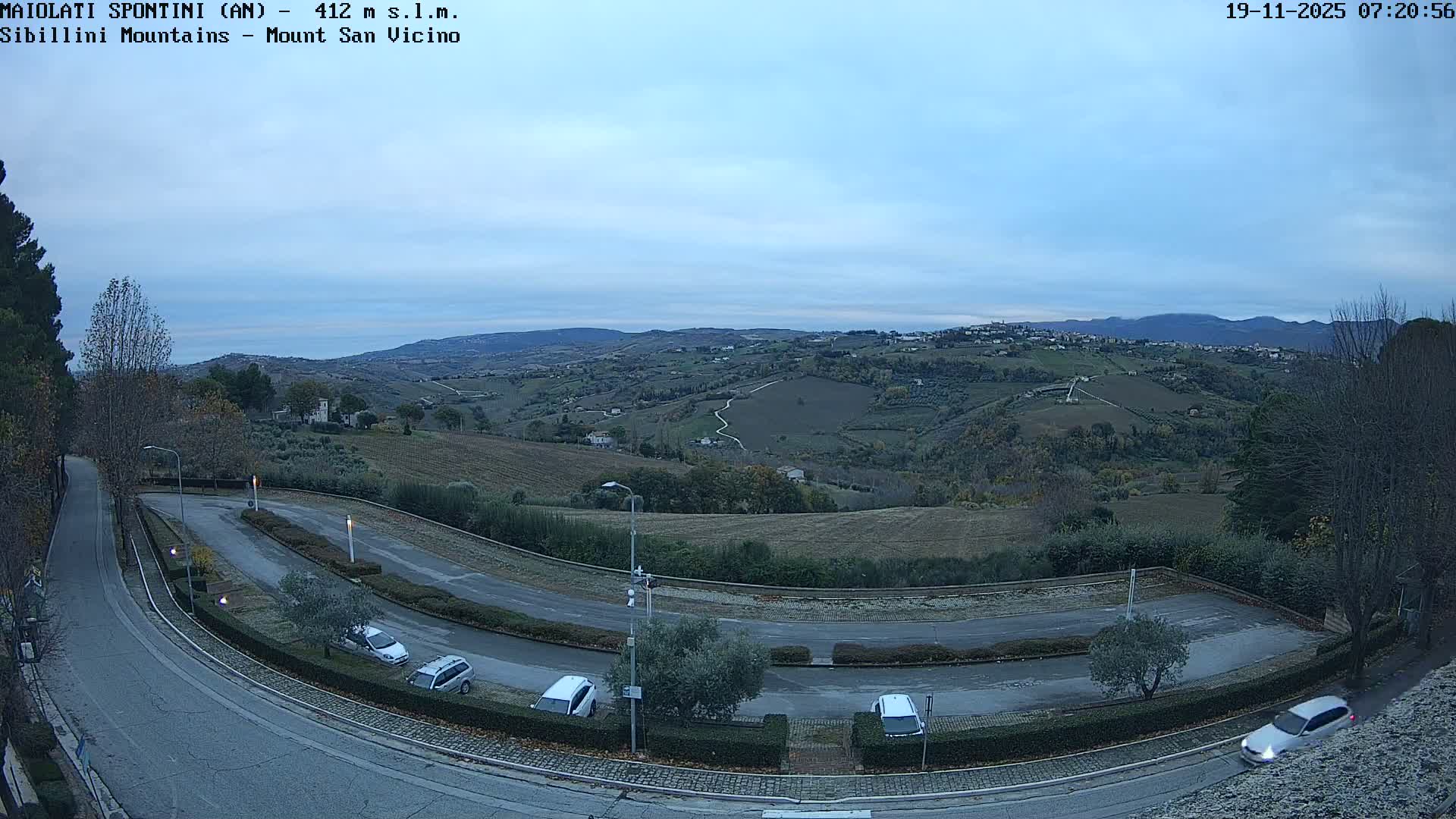 A wide-angle view captures a winding road with parked cars in the foreground, leading into a vast landscape of rolling, autumnal hills with fields, scattered trees, and distant hazy mountains under an overcast sky.