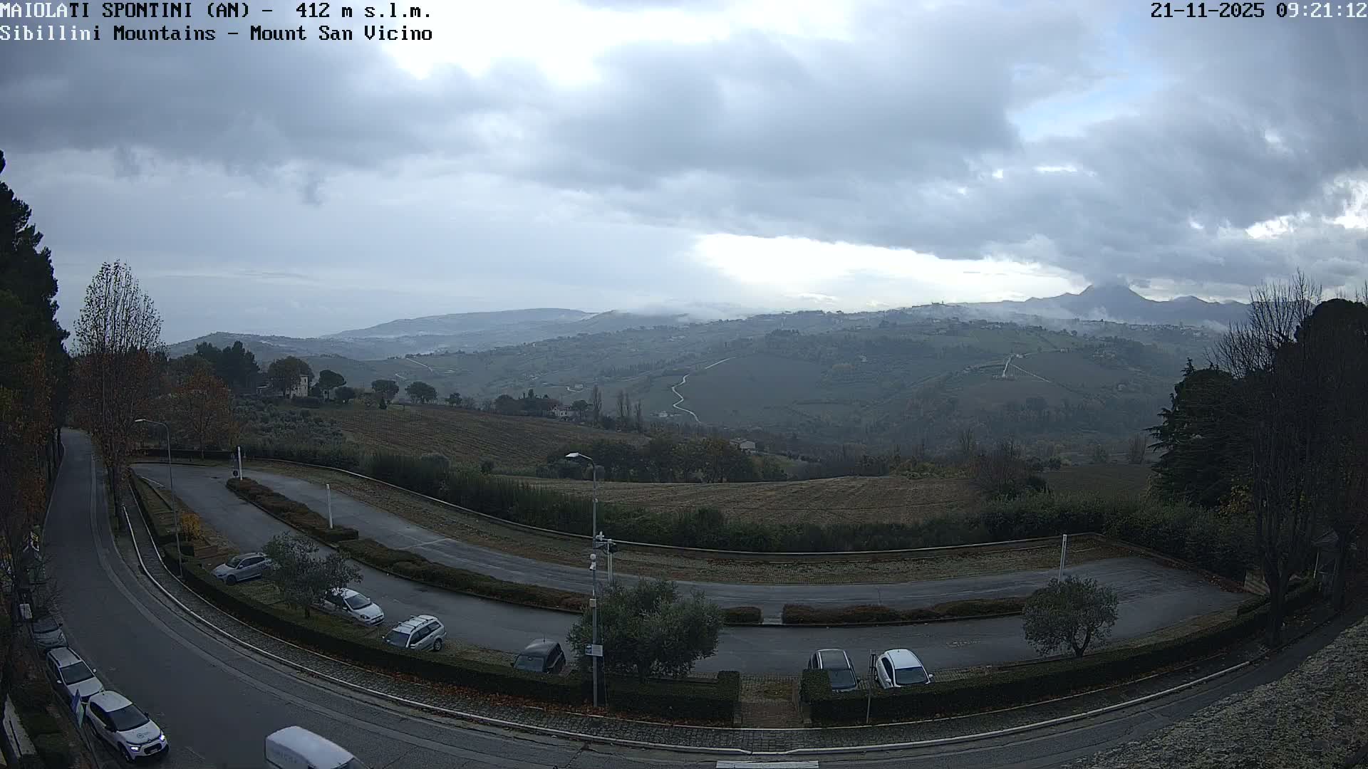 A wide view captures a winding road with parked cars and cultivated fields in the foreground, leading to rolling hills and distant mountains partially obscured by mist, all under an overcast and gloomy sky.
