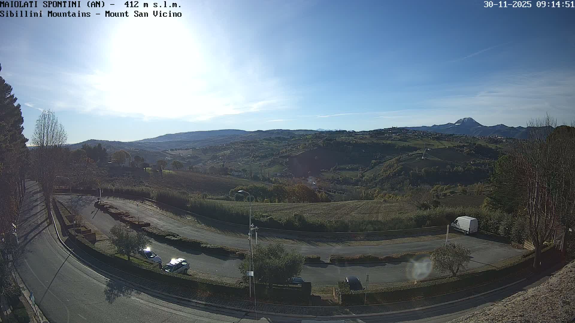 On a bright sunny day with a clear blue sky, a winding road in the foreground features several parked vehicles and leads into a sprawling panorama of rolling green and brown hills dotted with trees, small villages, and cultivated fields, all set against a backdrop of distant mountains, one prominent snow-capped peak.