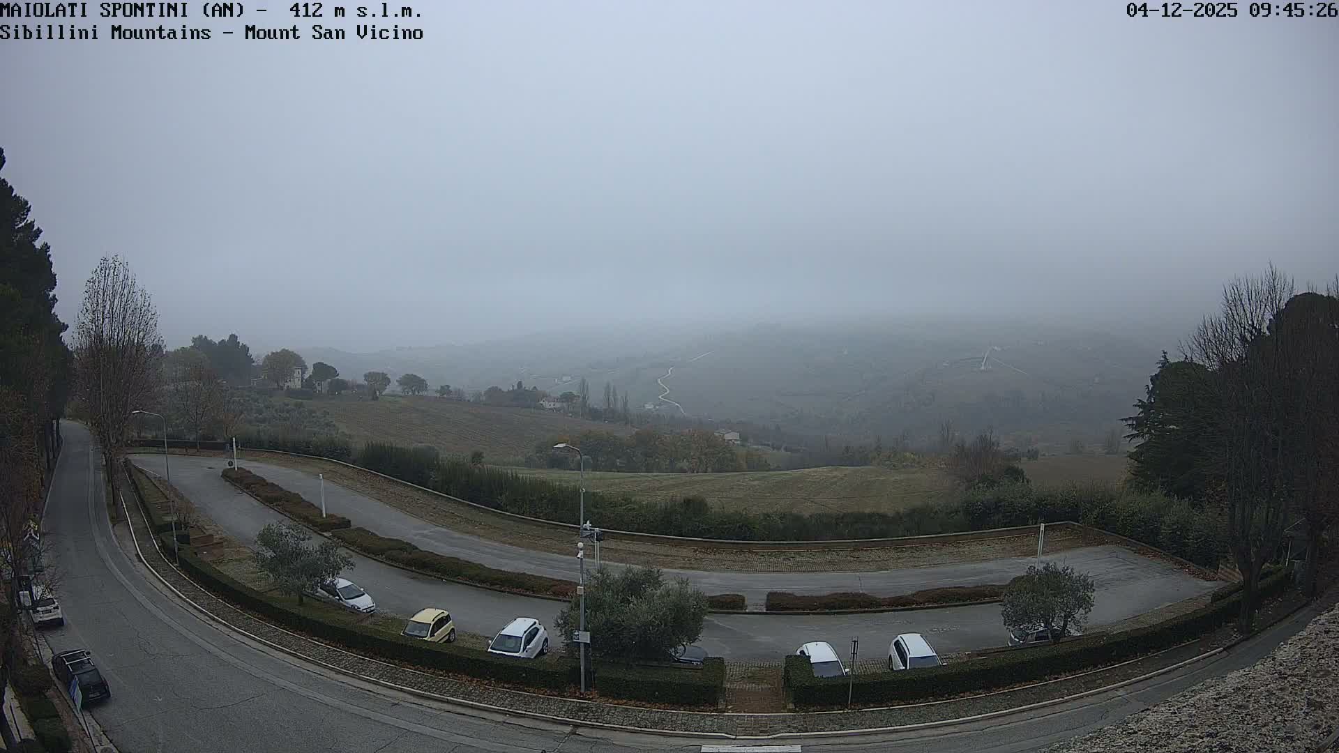 A foggy and overcast outdoor scene features a winding road with parked cars in the foreground, overlooking rolling hills and cultivated fields that fade into the distant mist.