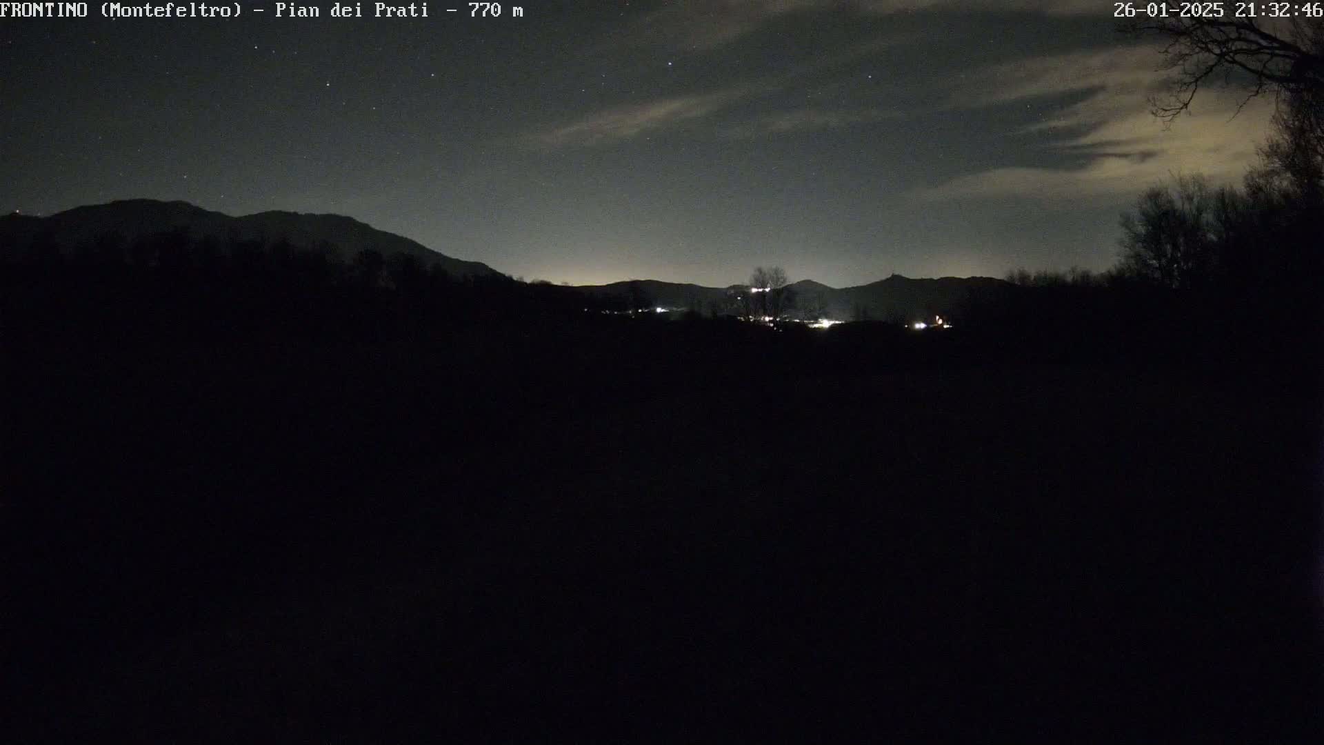 A nighttime view of a dark landscape with distant mountains under a slightly cloudy sky sprinkled with stars.