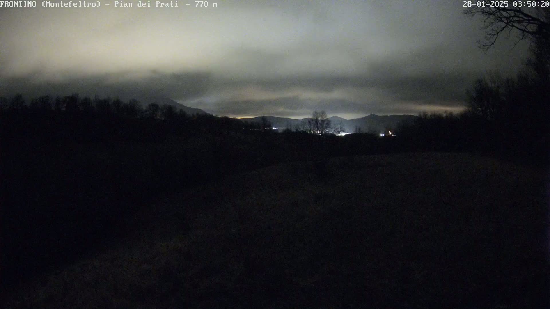 A nighttime view of a dark, overcast landscape showing distant lights from a town nestled among silhouetted hills and trees.