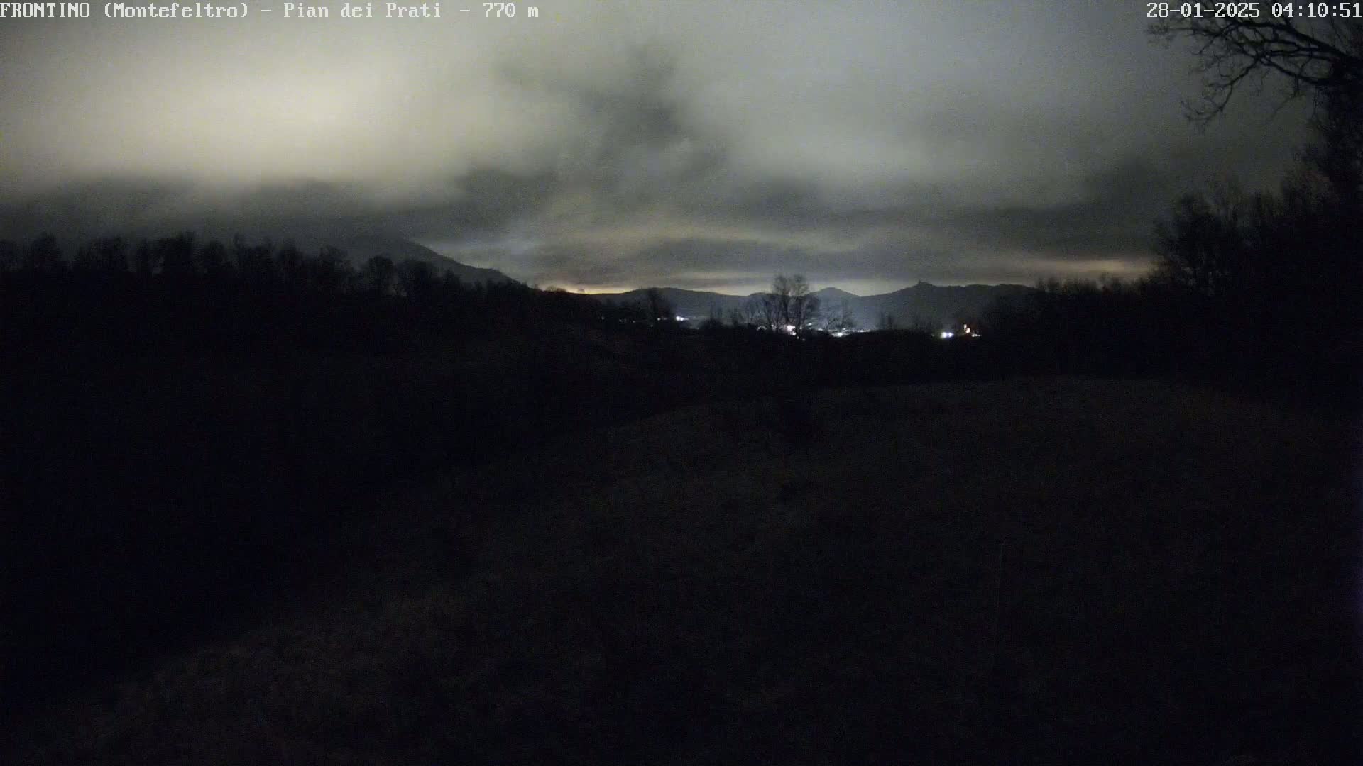 A nighttime view of dark, silhouetted trees and hills under a cloudy sky, with a few distant lights visible.
