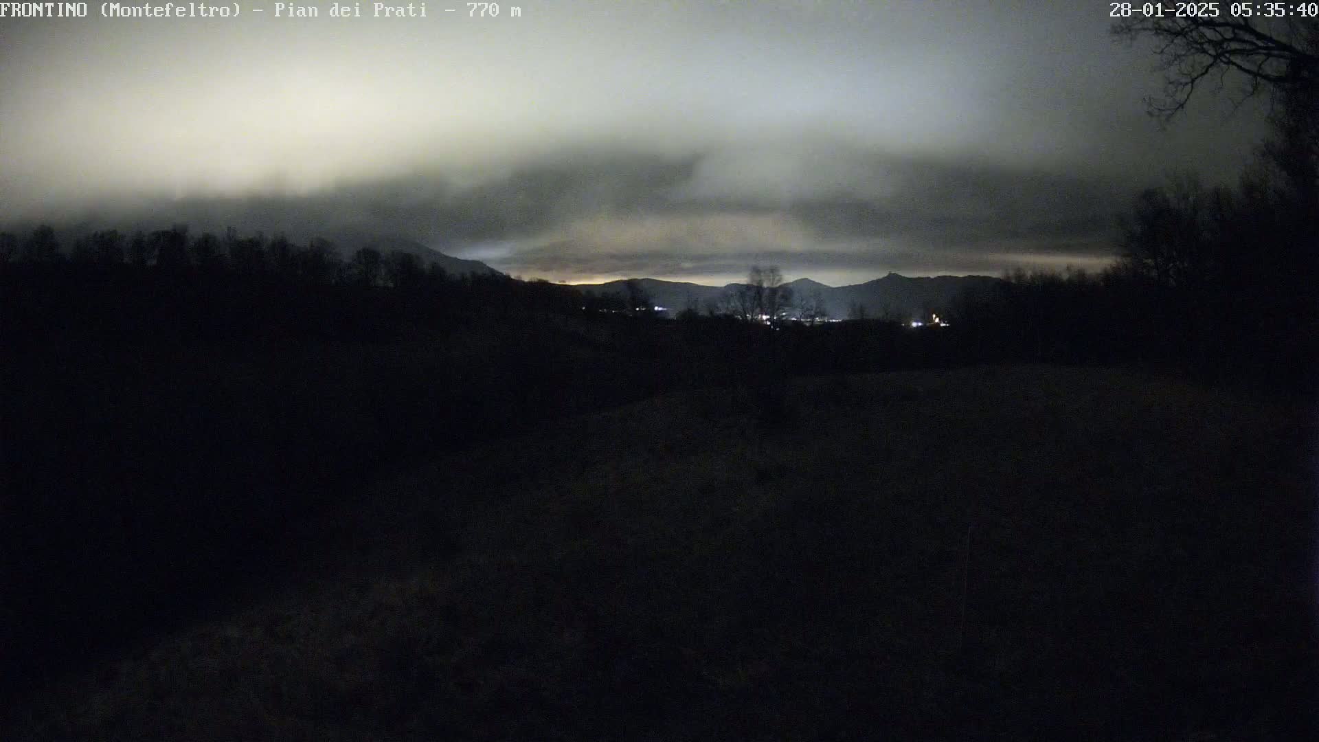 A dark, overcast night scene shows a silhouetted landscape of trees and distant mountains under a cloudy sky.