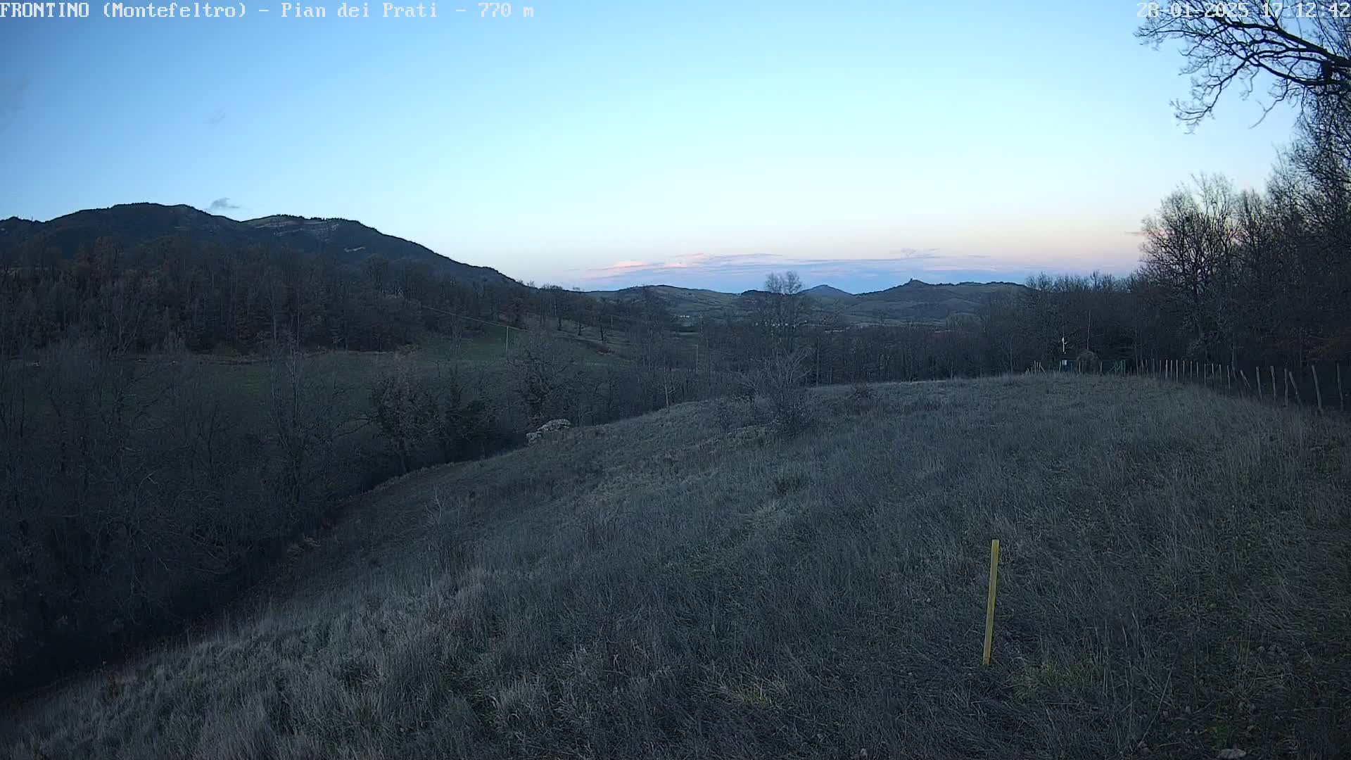A high-angle view shows a field of dry grass and leafless trees on a hillside overlooking a valley with more hills in the distance under a clear twilight sky.