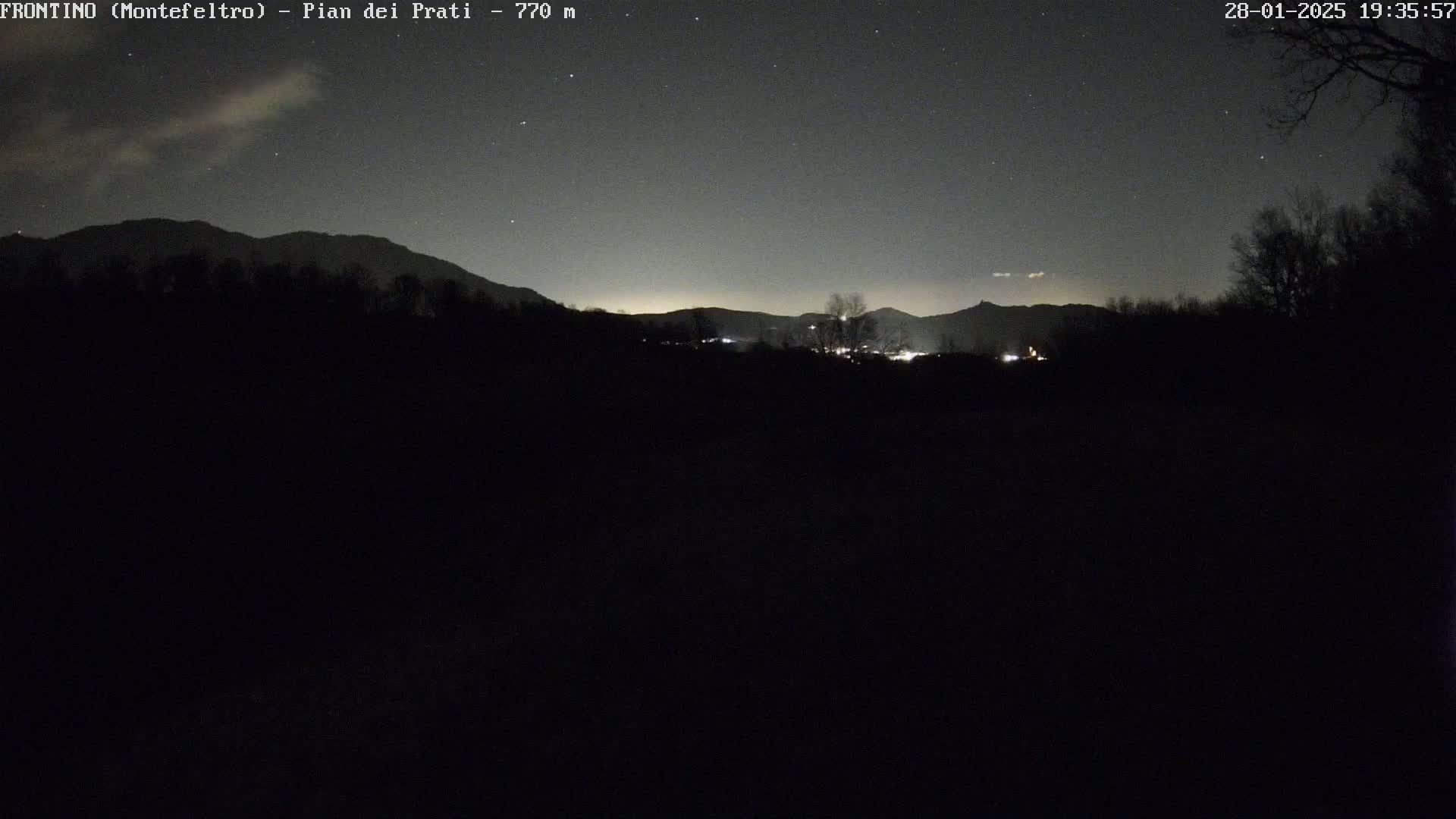 A nighttime, starry sky overlooks a dark, silhouetted landscape of mountains and trees, with faint lights visible in the distance.