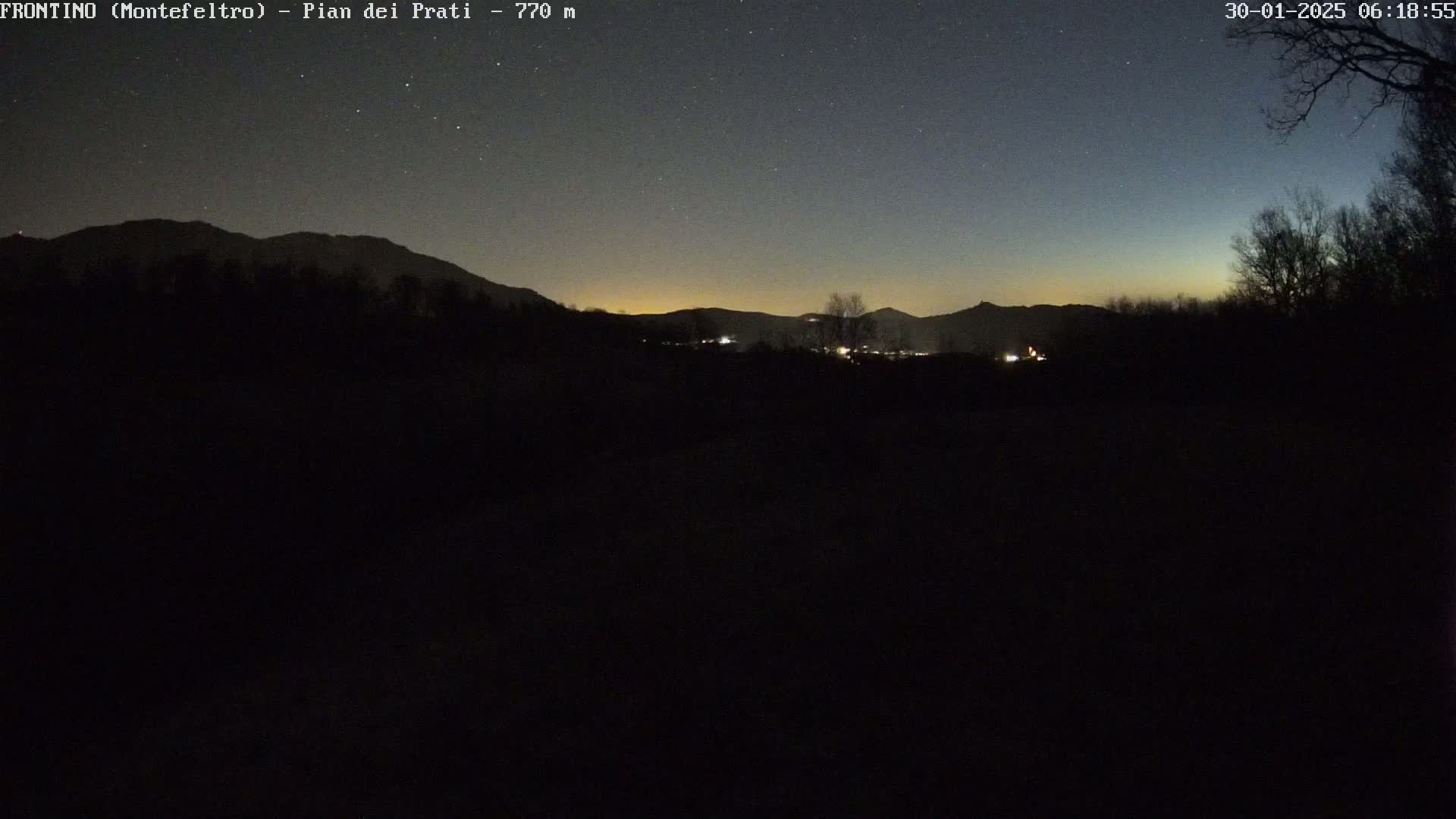 A dark, nighttime landscape shows a line of silhouetted mountains under a clear, starry sky with a faint, orange-tinged horizon.
