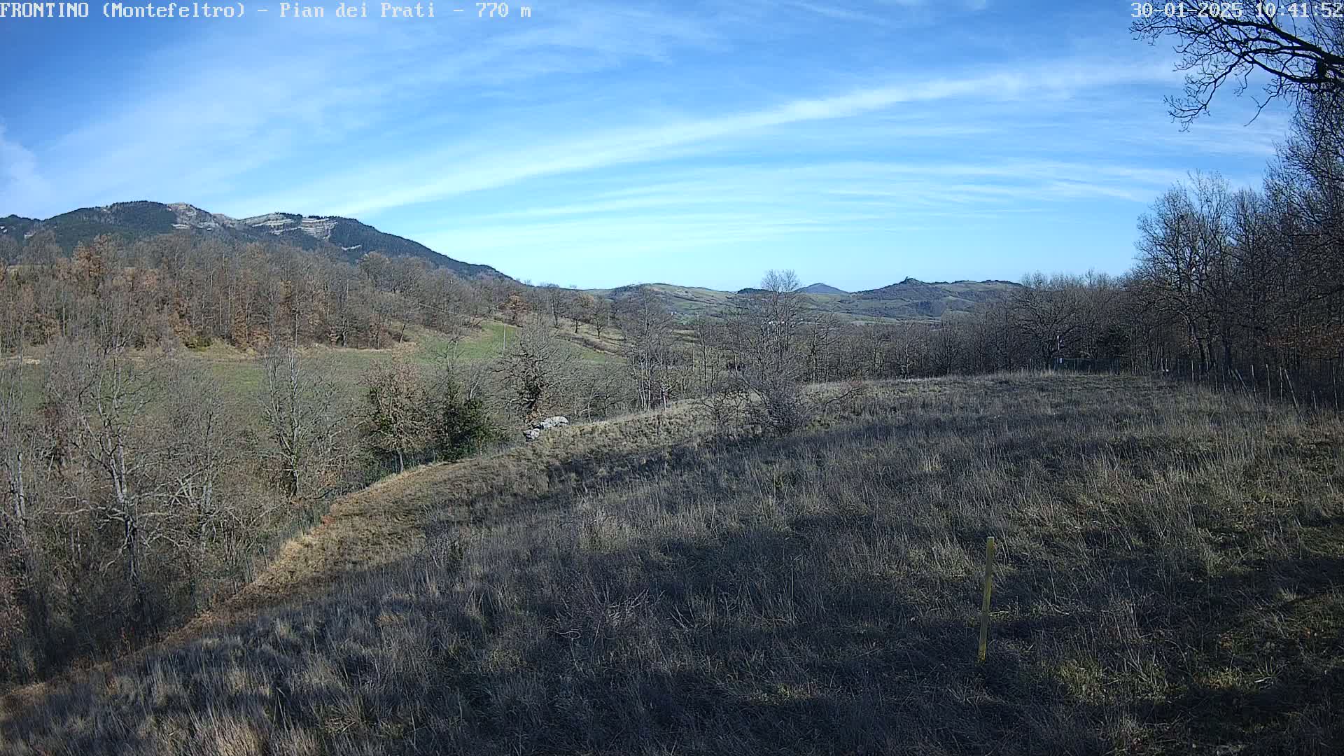 A mostly sunny, clear day reveals a sprawling landscape of dry, brown fields and leafless trees, nestled amidst rolling hills and mountains.