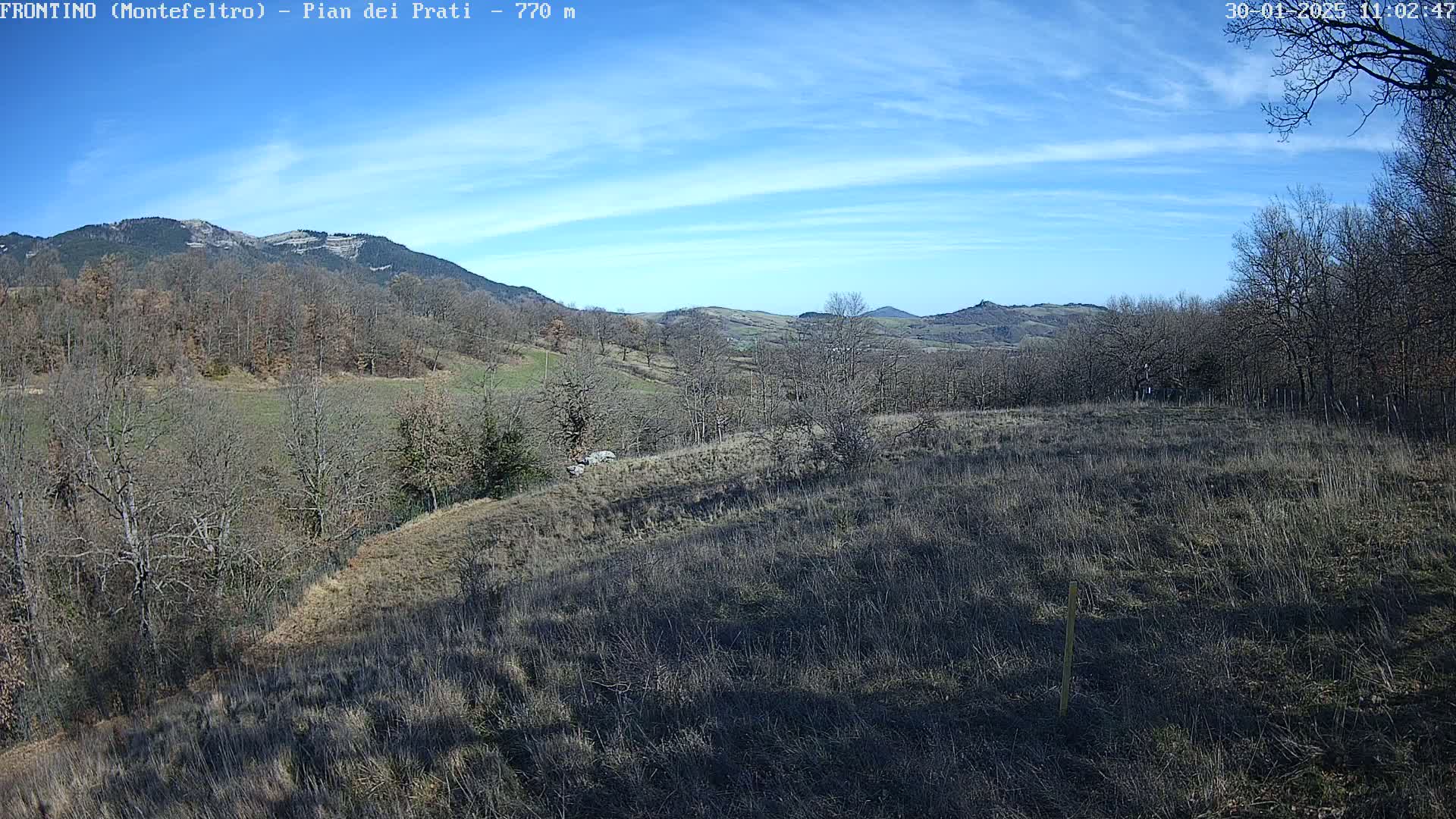 A mostly sunny day reveals a hilly landscape with leafless trees and dry, brown fields, extending to distant mountains under a pale blue sky.