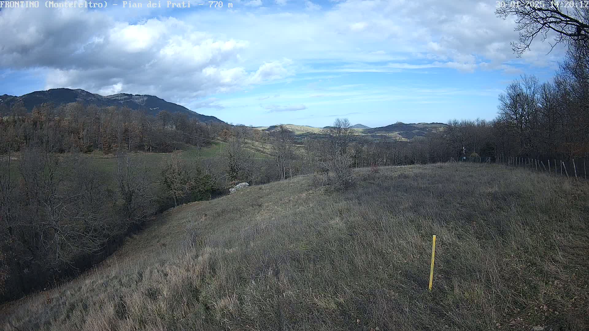 A view of a hilly landscape under a partly cloudy sky, showing bare winter trees and dry, brown fields.