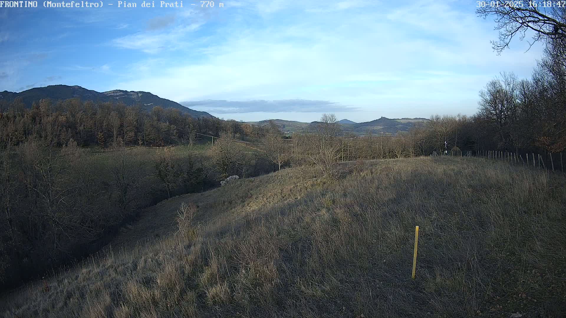 A partly cloudy sky overlooks a hilly landscape of leafless trees and dry, brown grass under a mostly clear sky.