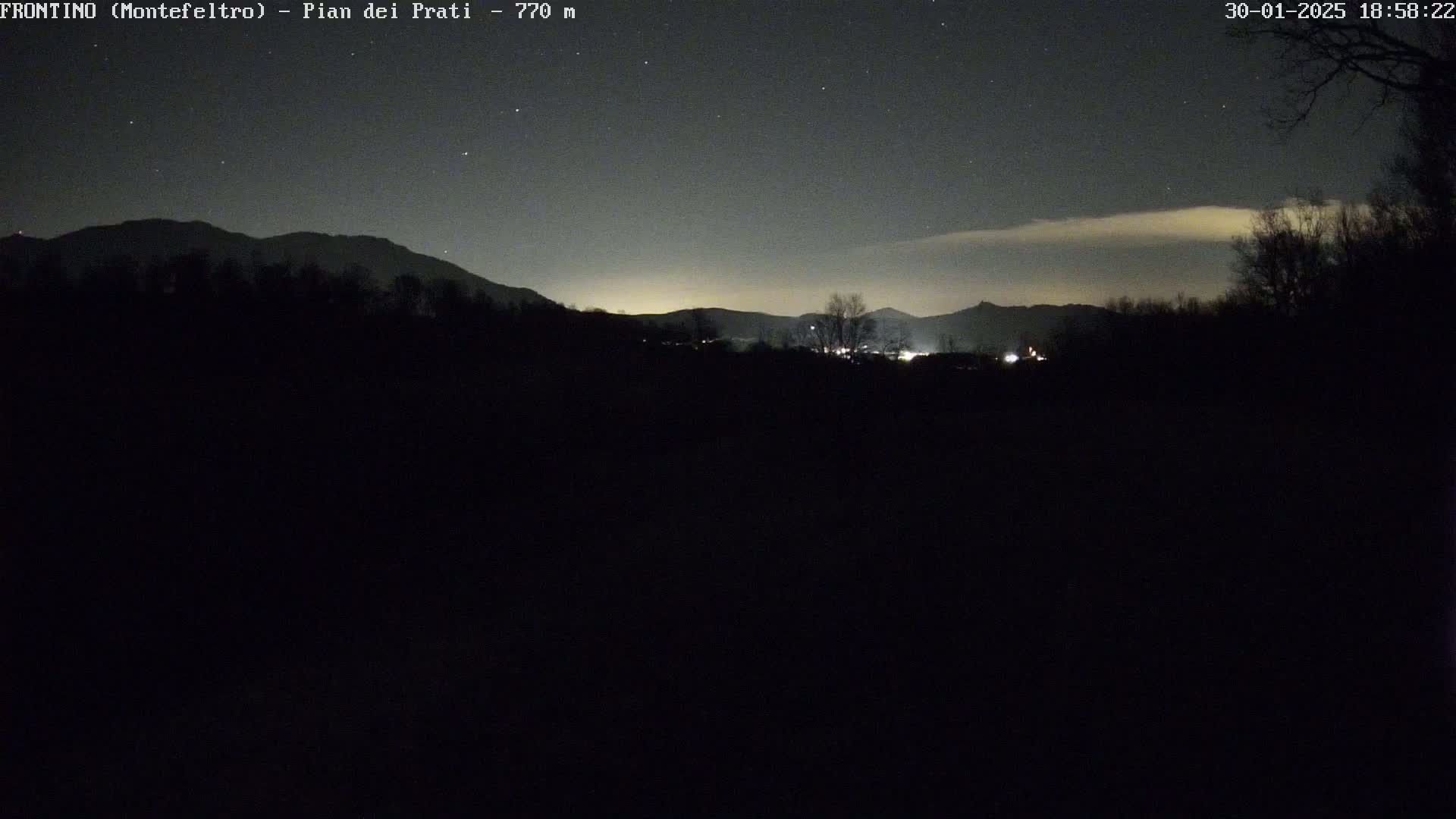 A nighttime view of dark silhouettes of mountains and trees under a mostly clear, starry sky with a faint band of clouds near the horizon.