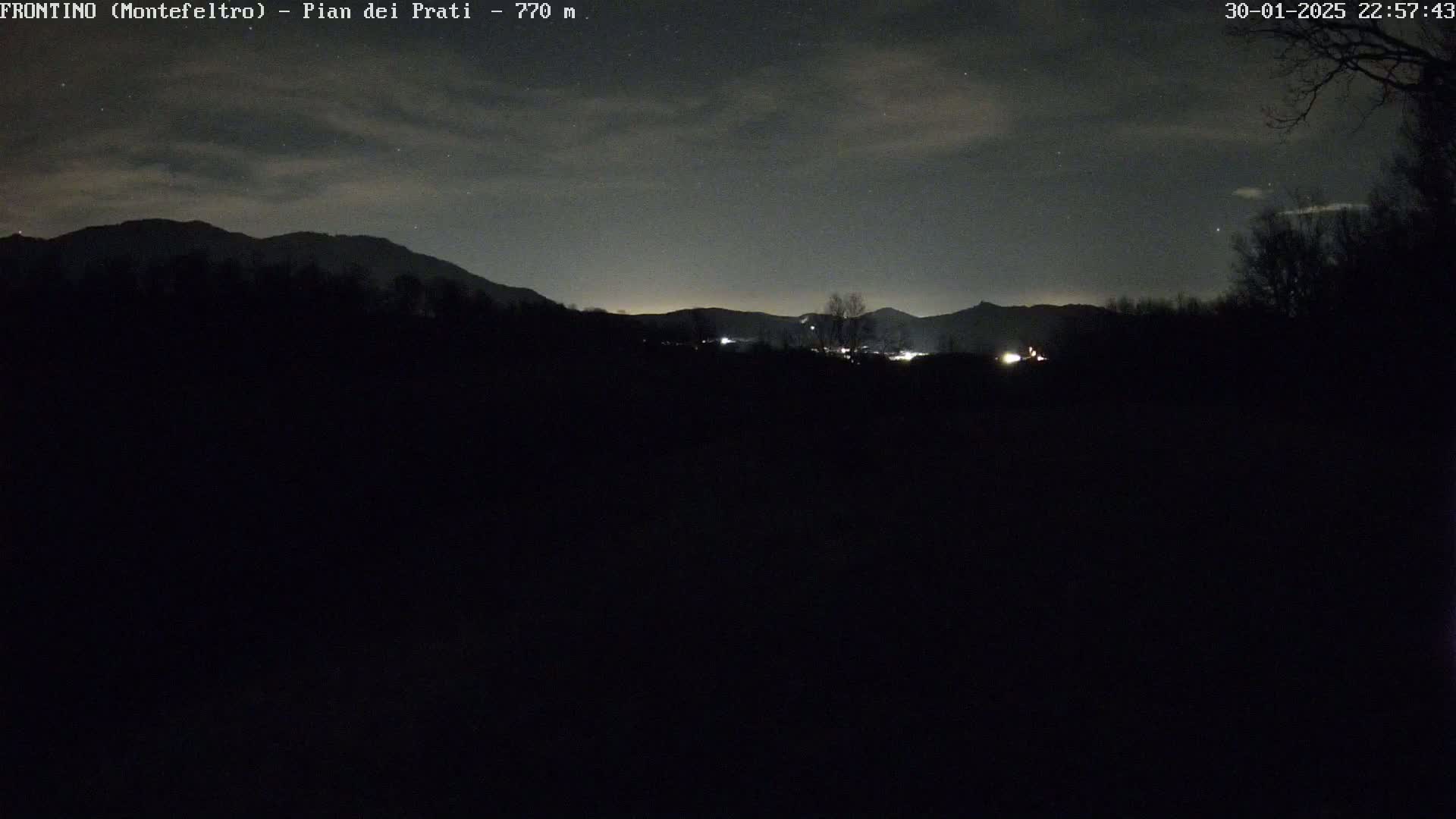 A nighttime view of dark silhouettes of mountains under a mostly clear sky with a few clouds and some stars visible.
