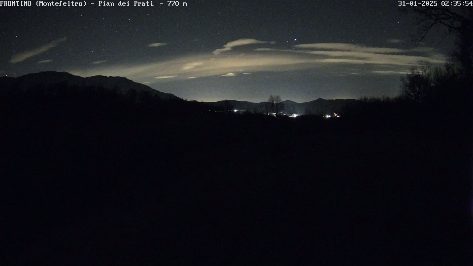 A nighttime landscape of dark silhouetted mountains and trees under a partly cloudy sky with visible stars.