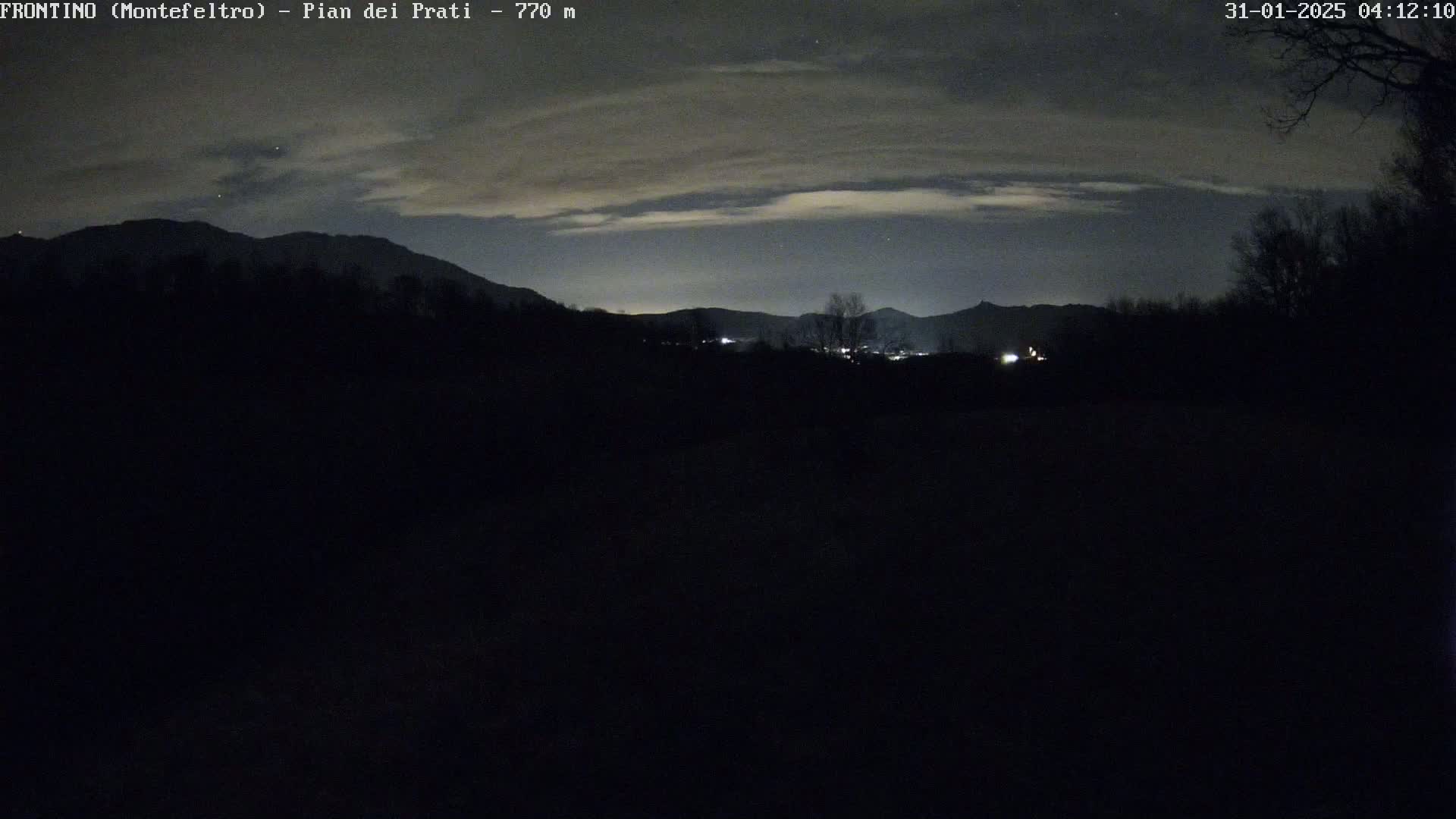 A nighttime view of a dark landscape with silhouetted mountains in the distance under a cloudy sky.