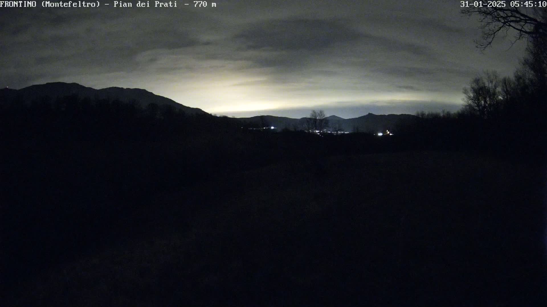 A dark, nighttime landscape shows silhouetted mountains under a cloudy, light-grey sky.