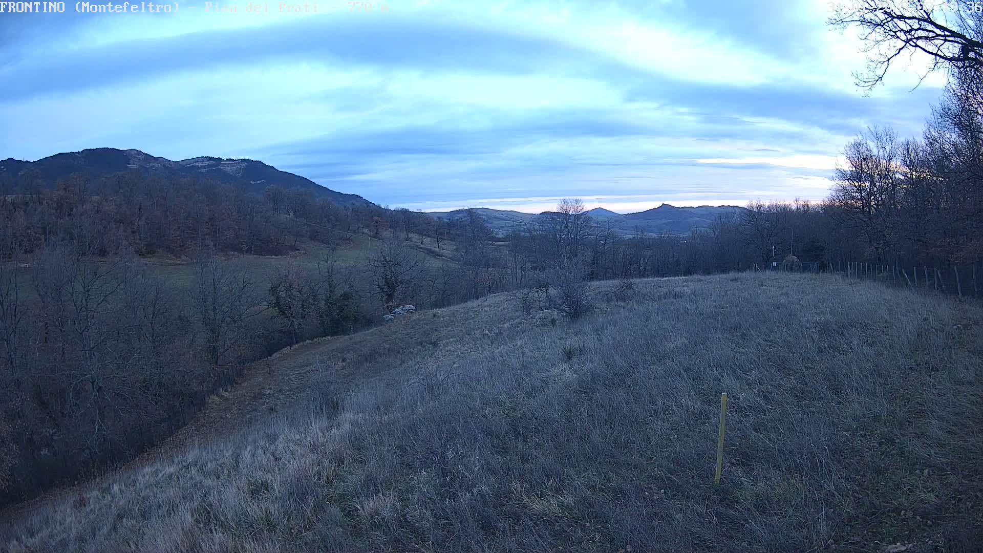 A view of a hilly landscape under a partly cloudy sky shows distant mountains and a foreground of dry, brown grass.