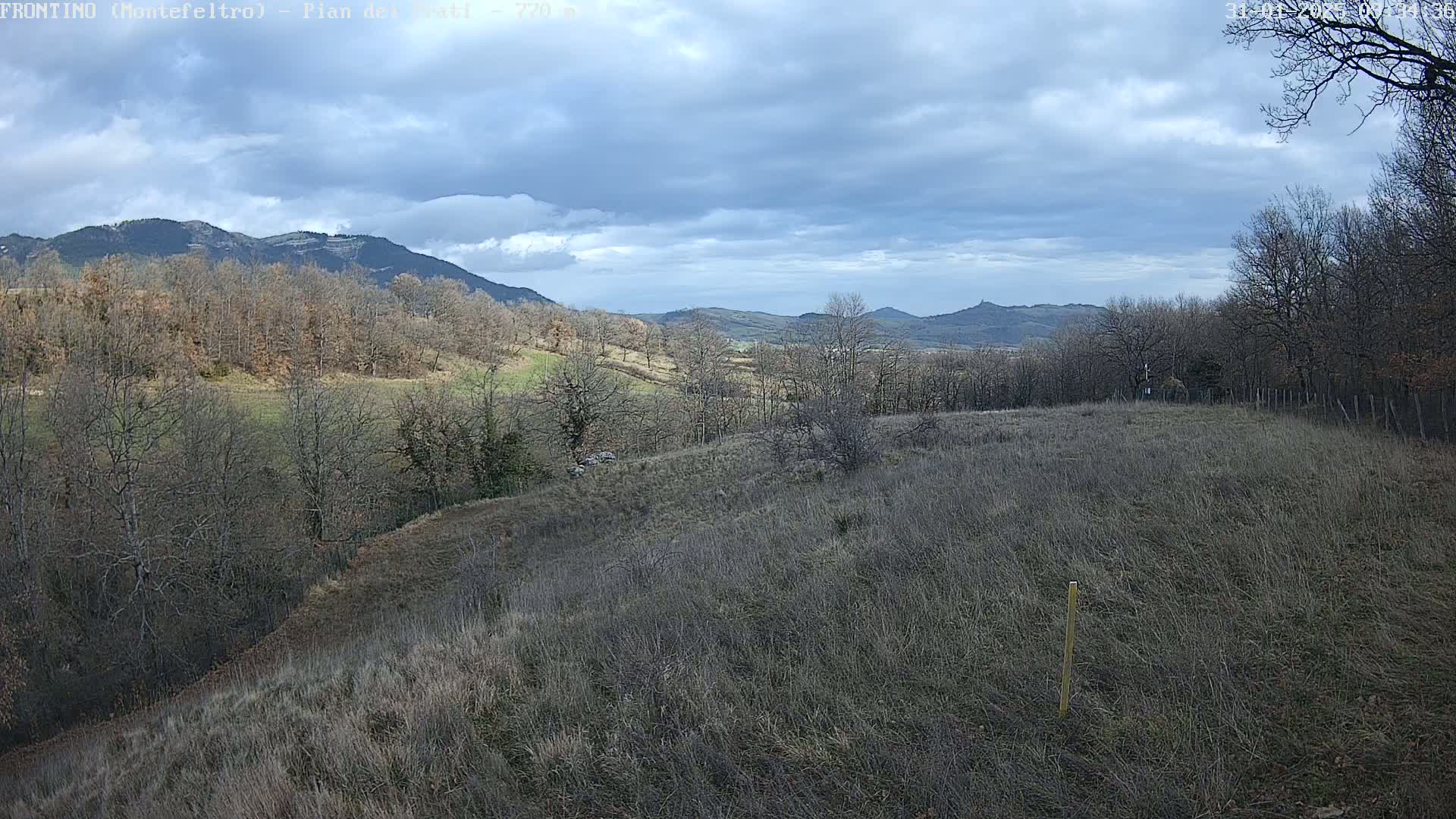 A sprawling, dry field sits atop a hill overlooking a valley of leafless trees and distant mountains under a partly cloudy sky.