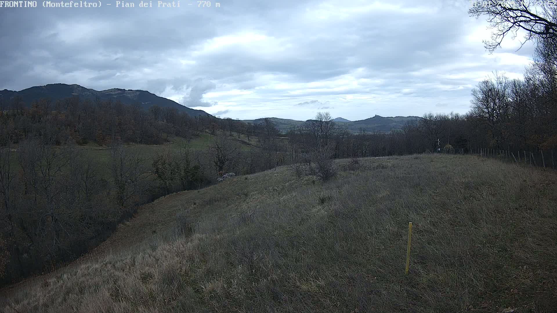 A view of a hilly landscape under a cloudy sky, showing barren, brown fields and leafless trees.