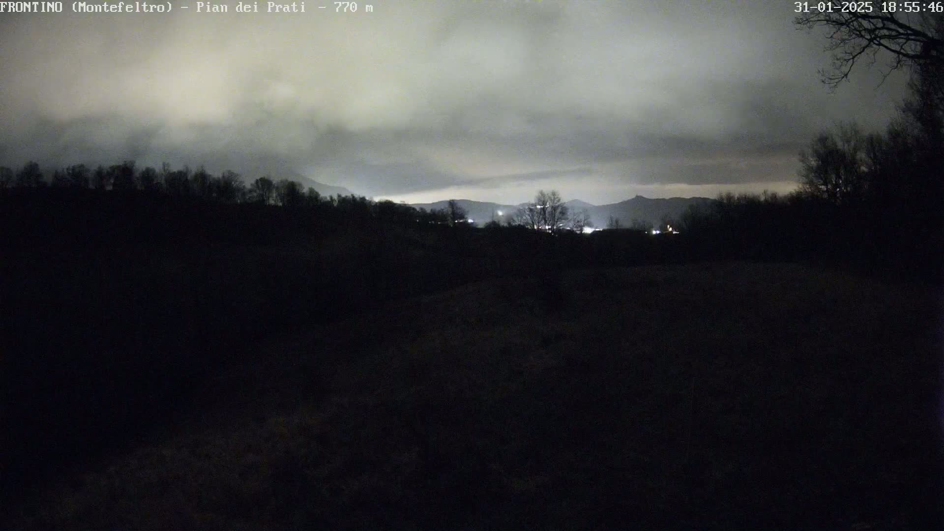 A nighttime view of a dark, silhouetted landscape under a cloudy sky, with faint lights visible in the distance.