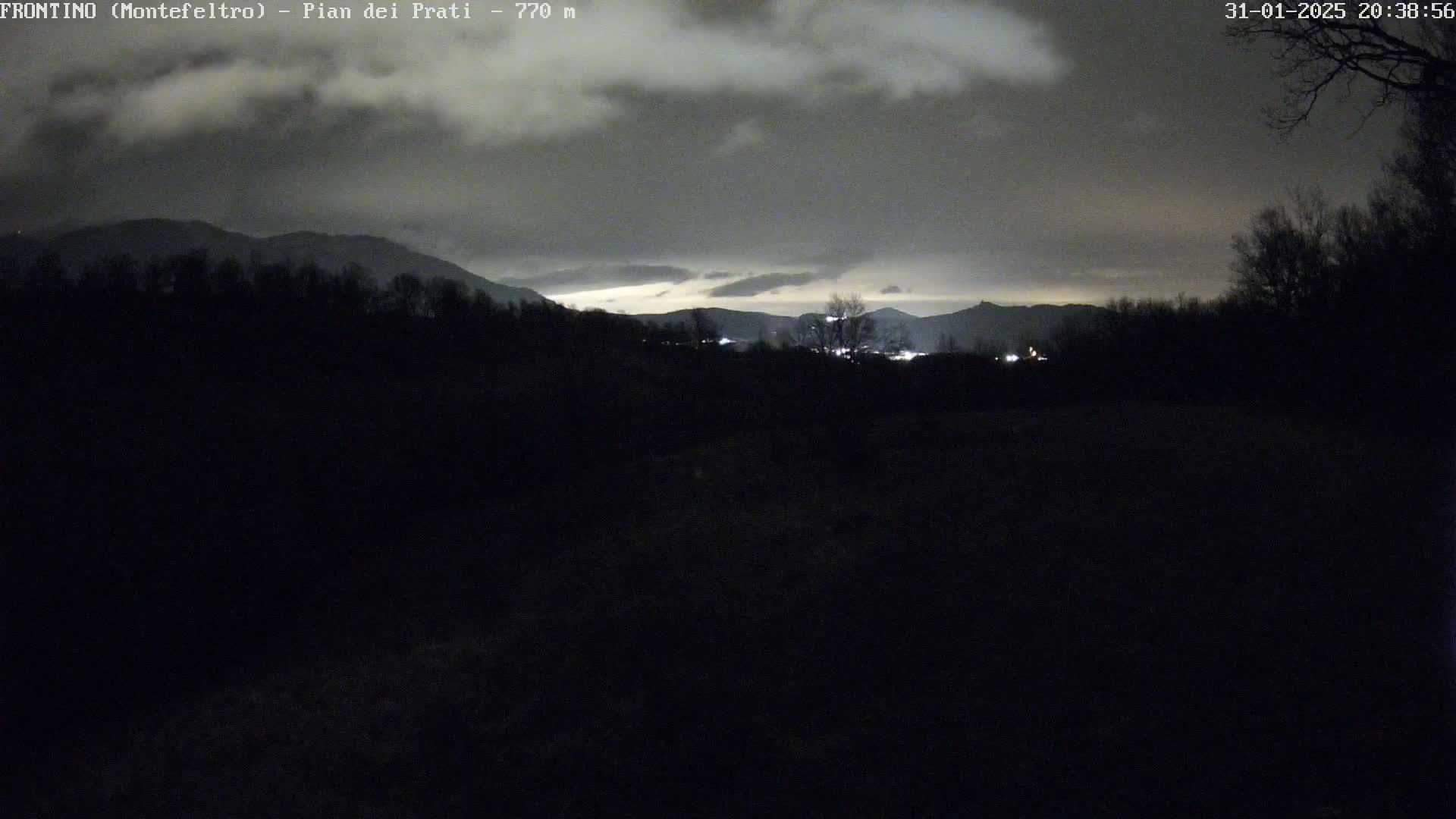 A nighttime view of dark, silhouetted hills and trees under a cloudy sky, with a faint illumination on the horizon.
