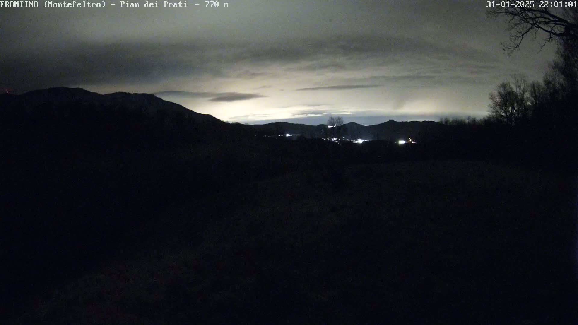 A dark, nighttime landscape shows silhouetted hills and a few distant lights under a cloudy sky.