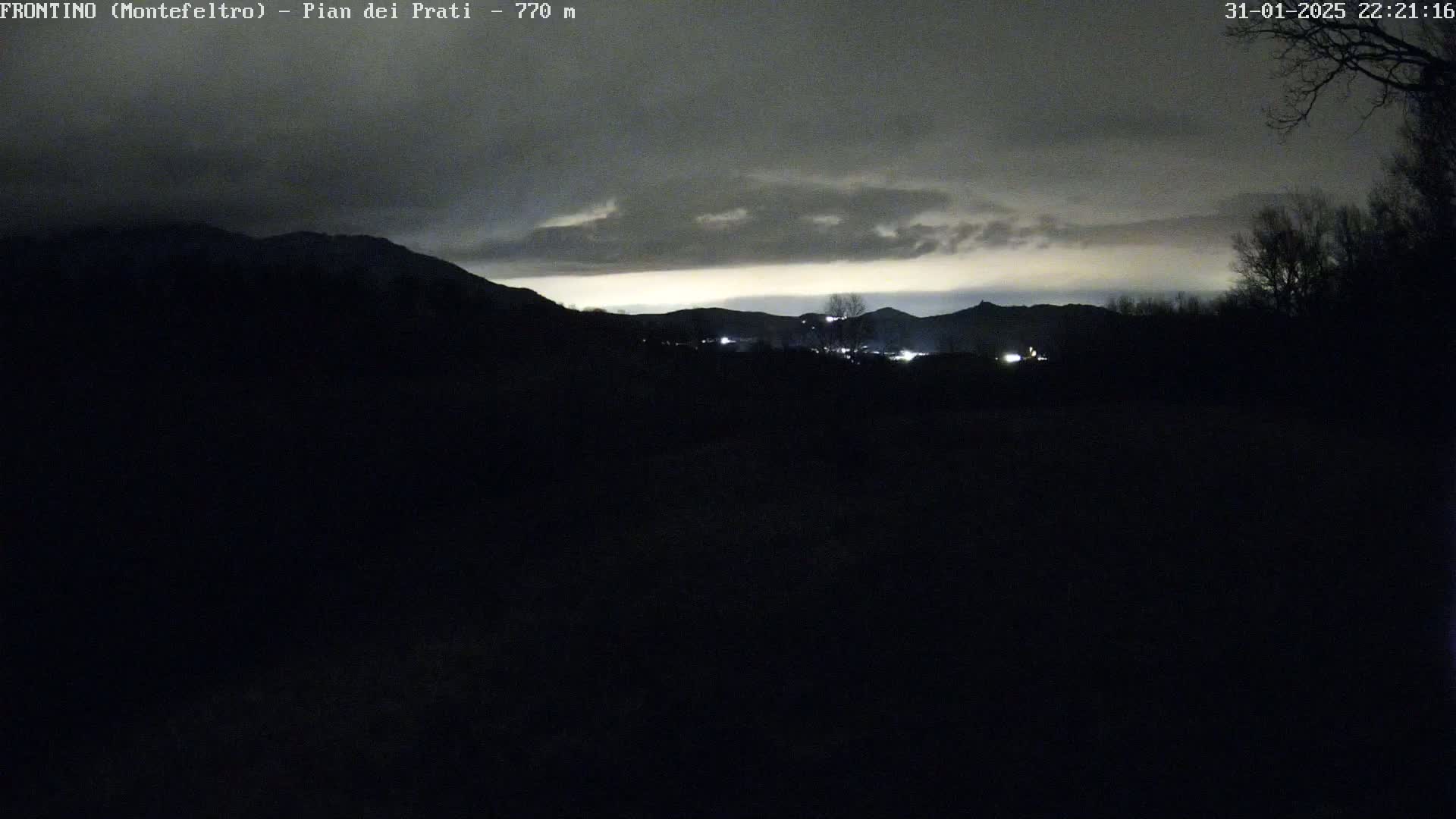 A nighttime landscape shows dark, silhouetted mountains and trees under a cloudy sky with a faint, light horizon.