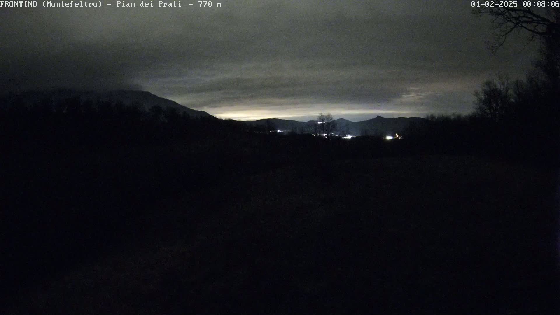 A nighttime, overcast view of a dark landscape with distant, faintly lit hills and mountains.