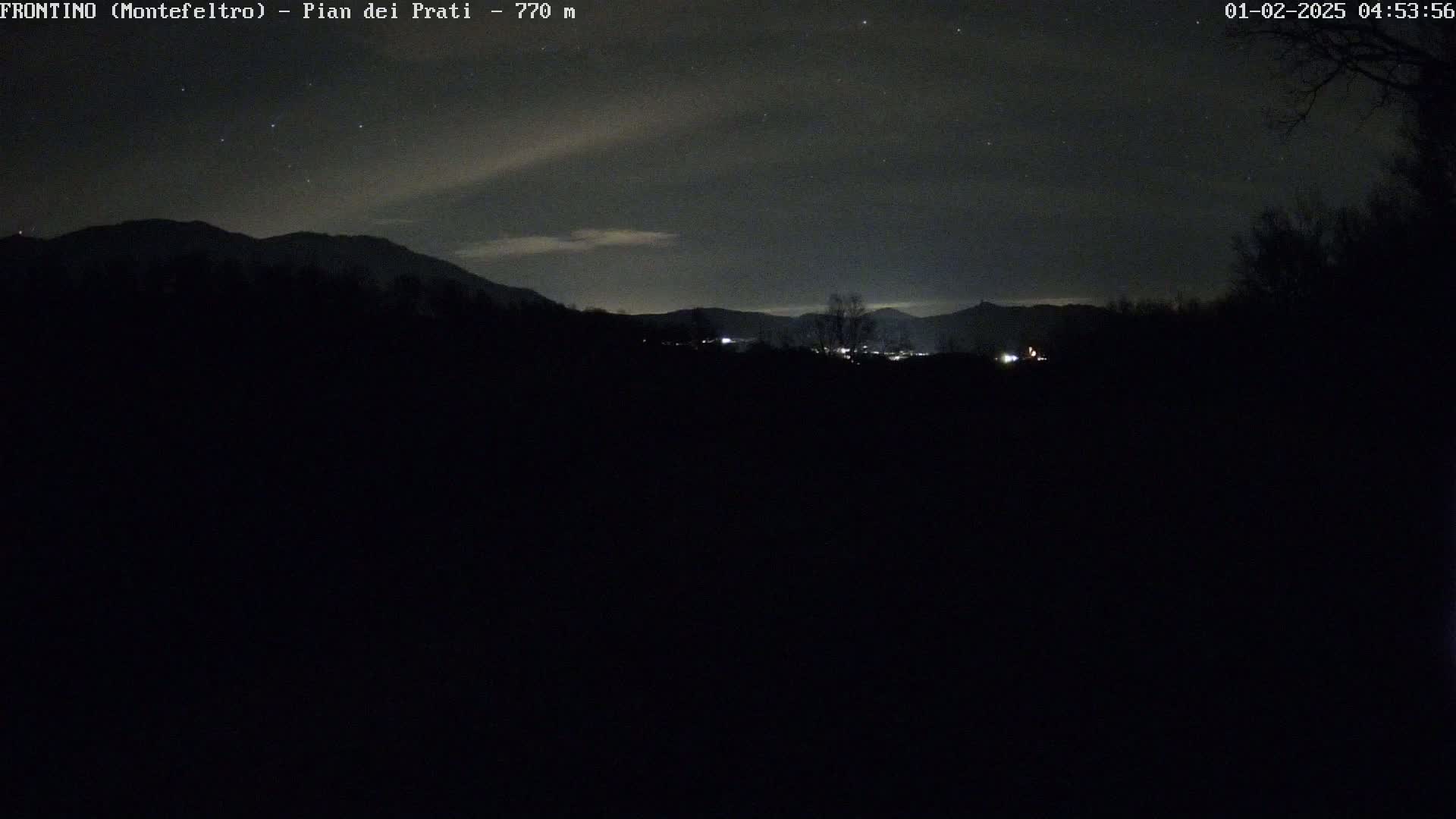 A nighttime view of a dark landscape with distant mountains under a partly cloudy sky dotted with stars.
