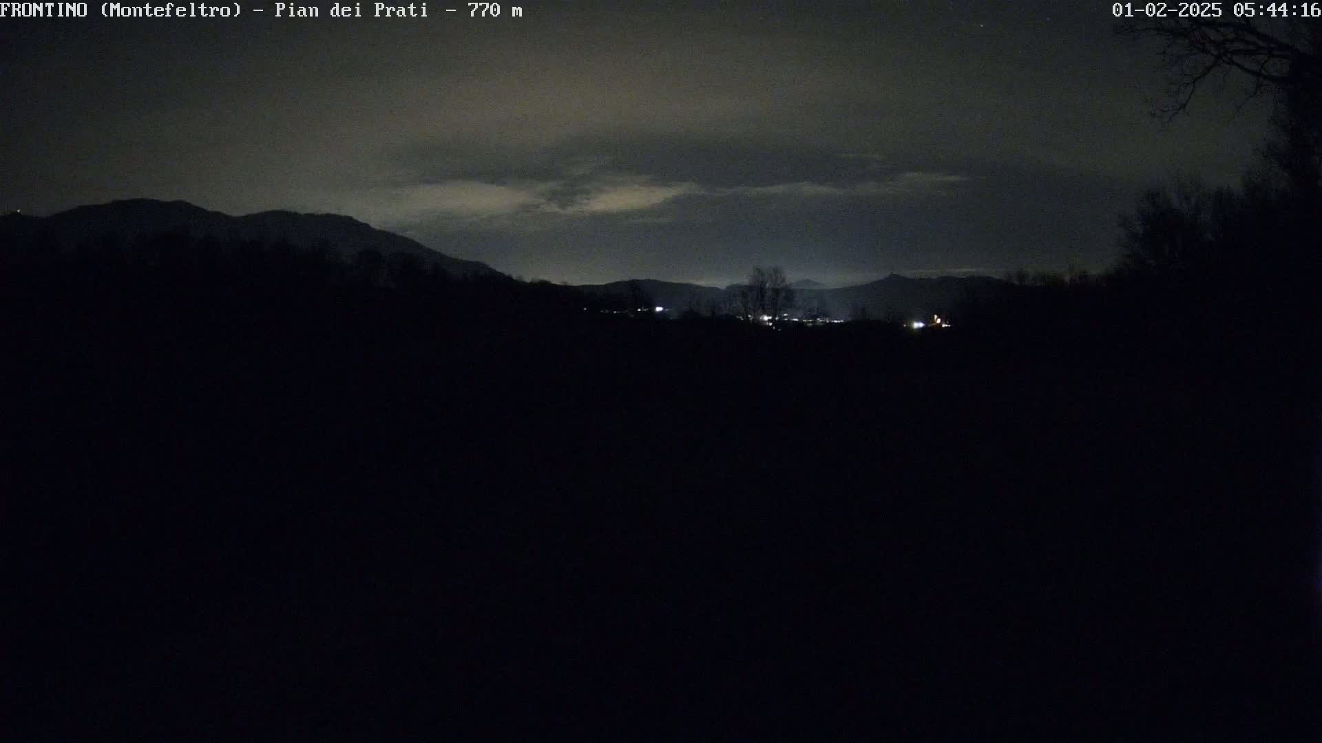 A dark, nighttime landscape shows distant, dimly lit buildings nestled amongst silhouetted mountains under a cloudy sky.