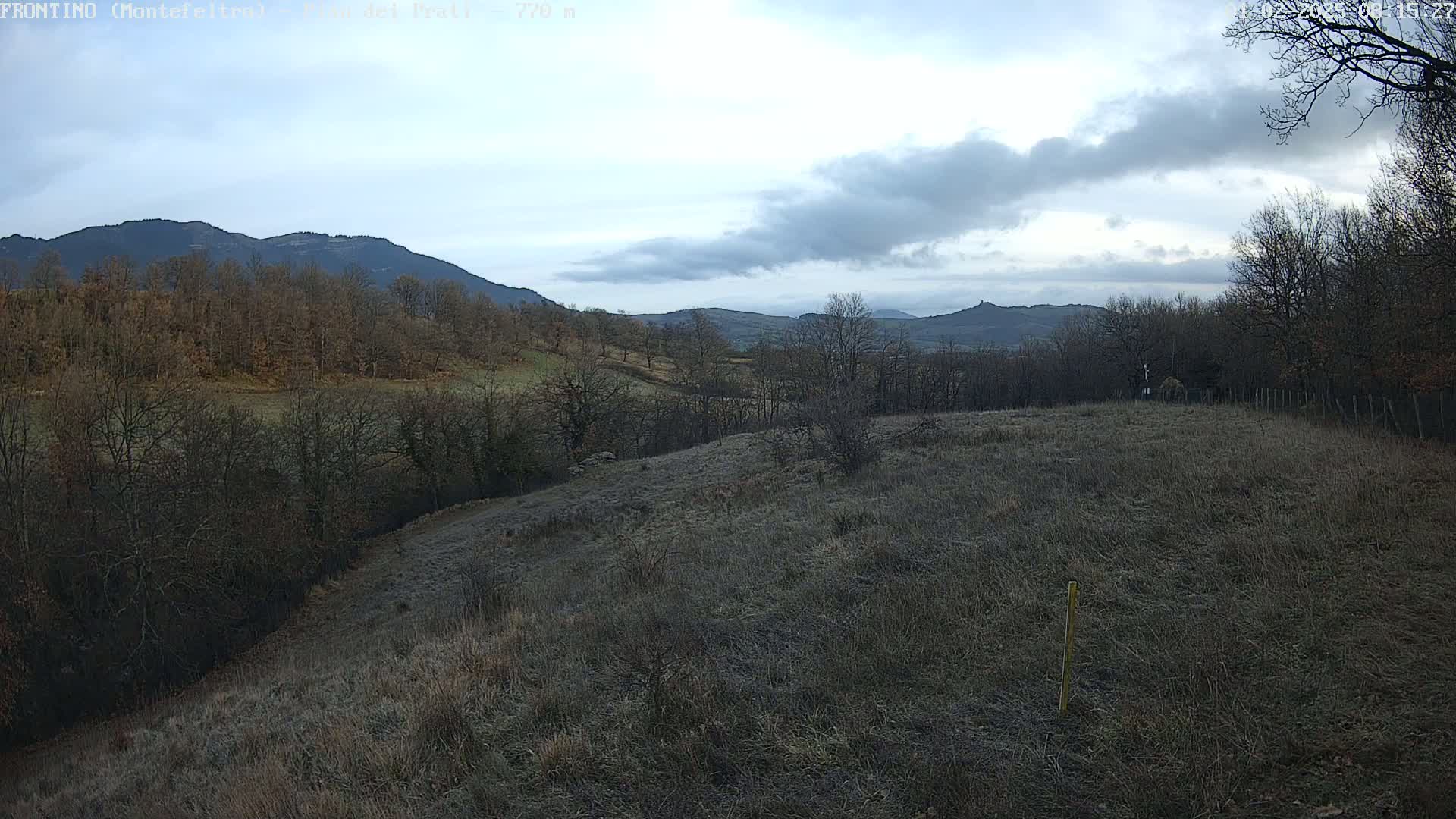 A mostly-clear, cool day reveals a hilly landscape with leafless trees and frost-covered fields, overlooking distant mountains.