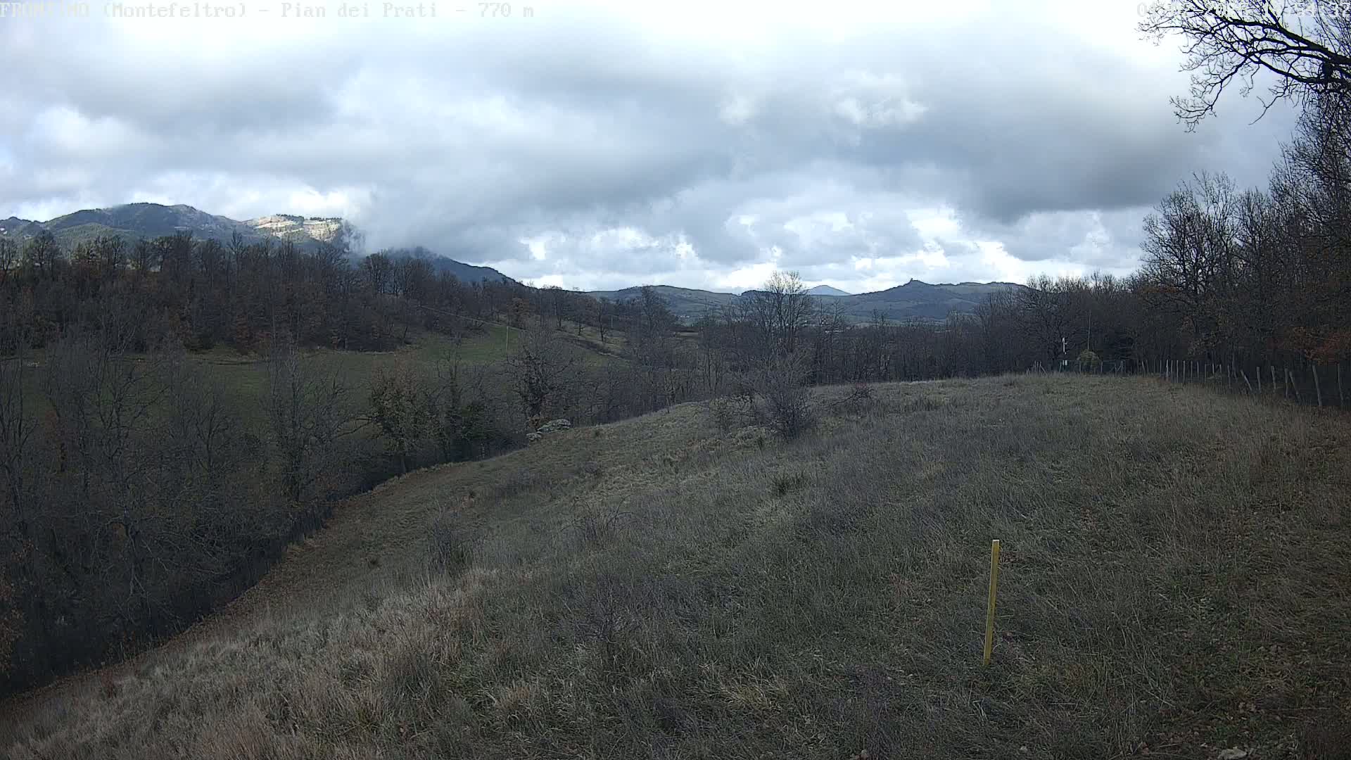 A mostly cloudy day reveals a hilly landscape with bare trees and dry, brown grass, and distant mountains under a gray sky.