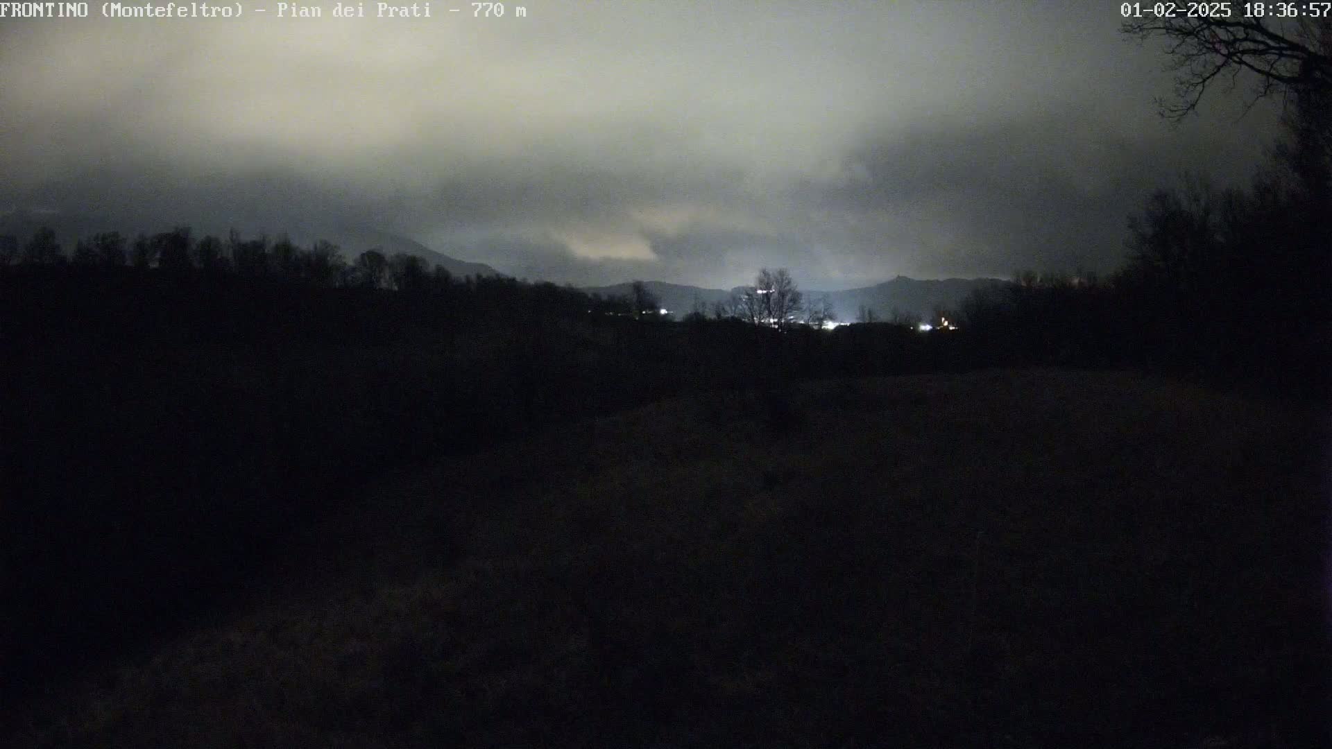 A dark, nighttime landscape shows a distant line of lights nestled between dark hills under a cloudy sky.