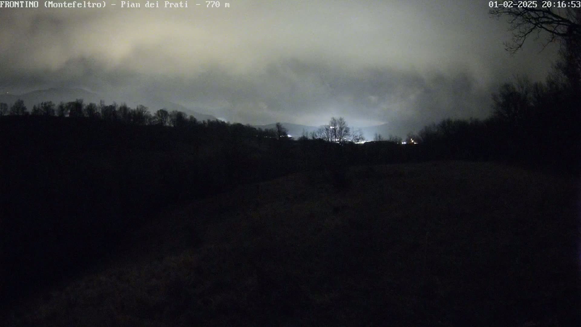 A nighttime view of a dark, hilly landscape under a cloudy sky, with faint lights visible in the distance.